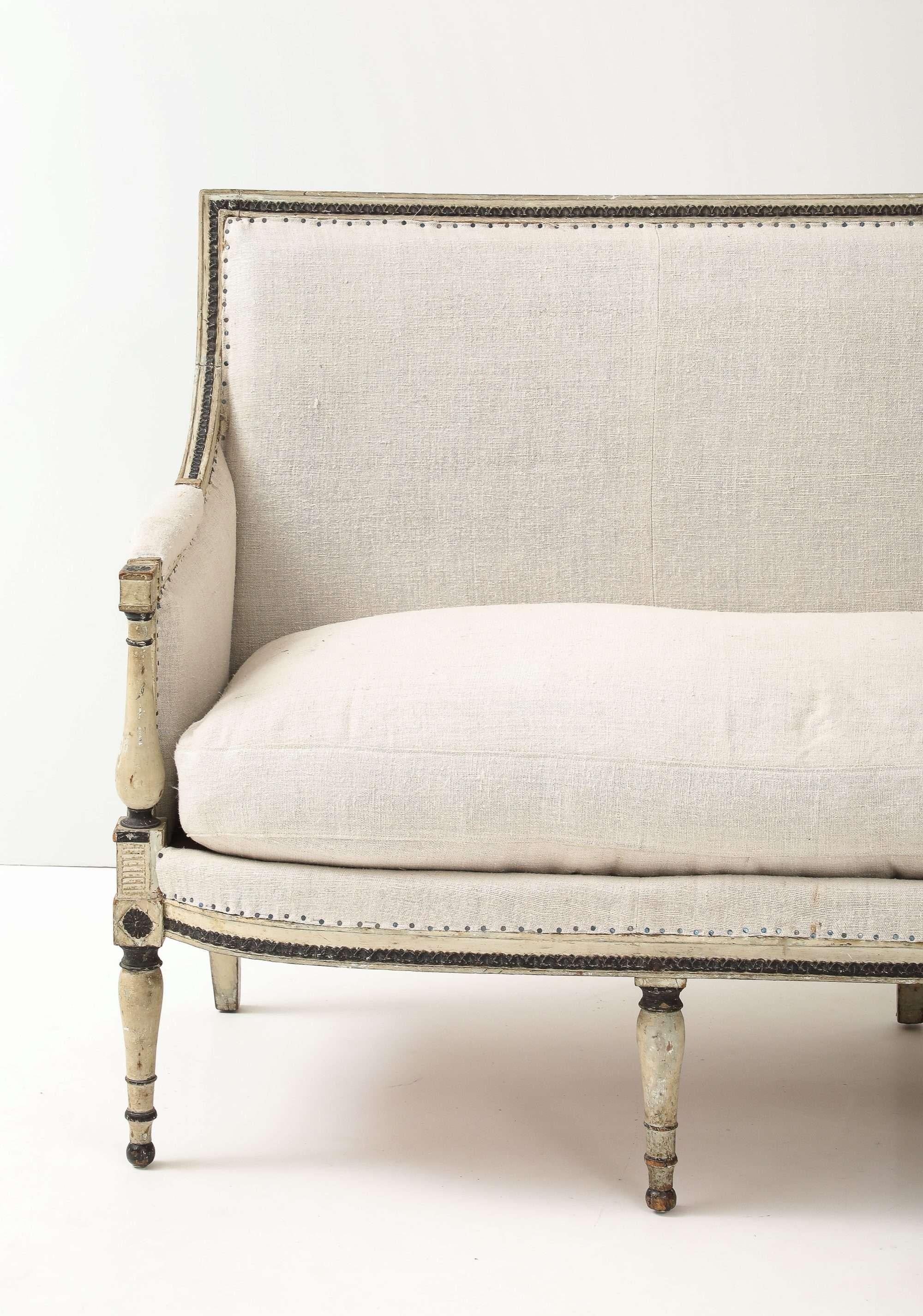 An elegant French settee retaining its original pale white with black accents painted finish and bronze nail-head trim. The straight back, slender arms, and six fluted tapering legs with rosette blocks showcase classic Louis XVI carving, while fresh