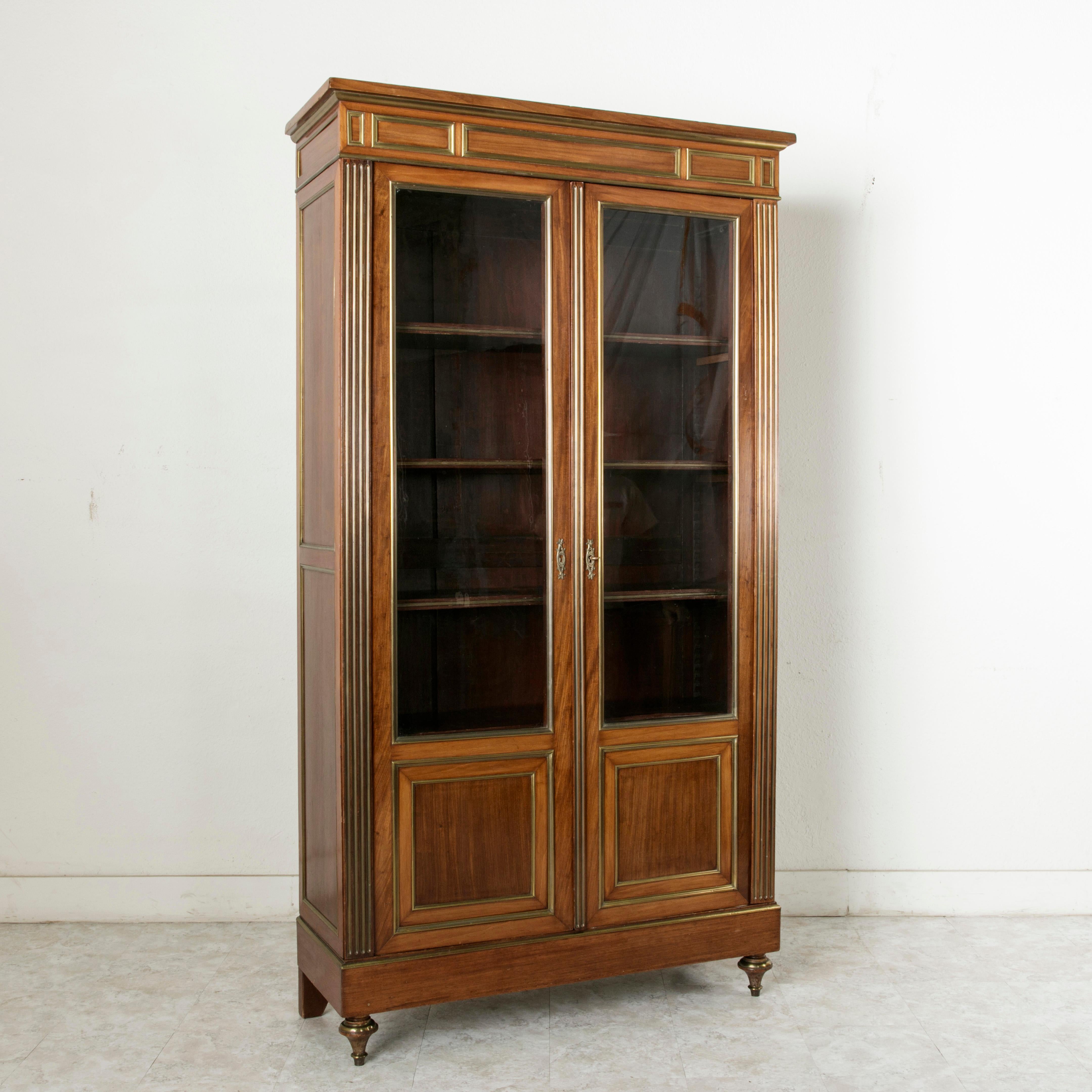 This striking 19th century Louis XVI style mahogany bibliotheque or bookcase is finished with stunning bronze banding. Its corners feature fluting with inset bronze filets. Both doors have their original blown glass, evidenced by waves and an