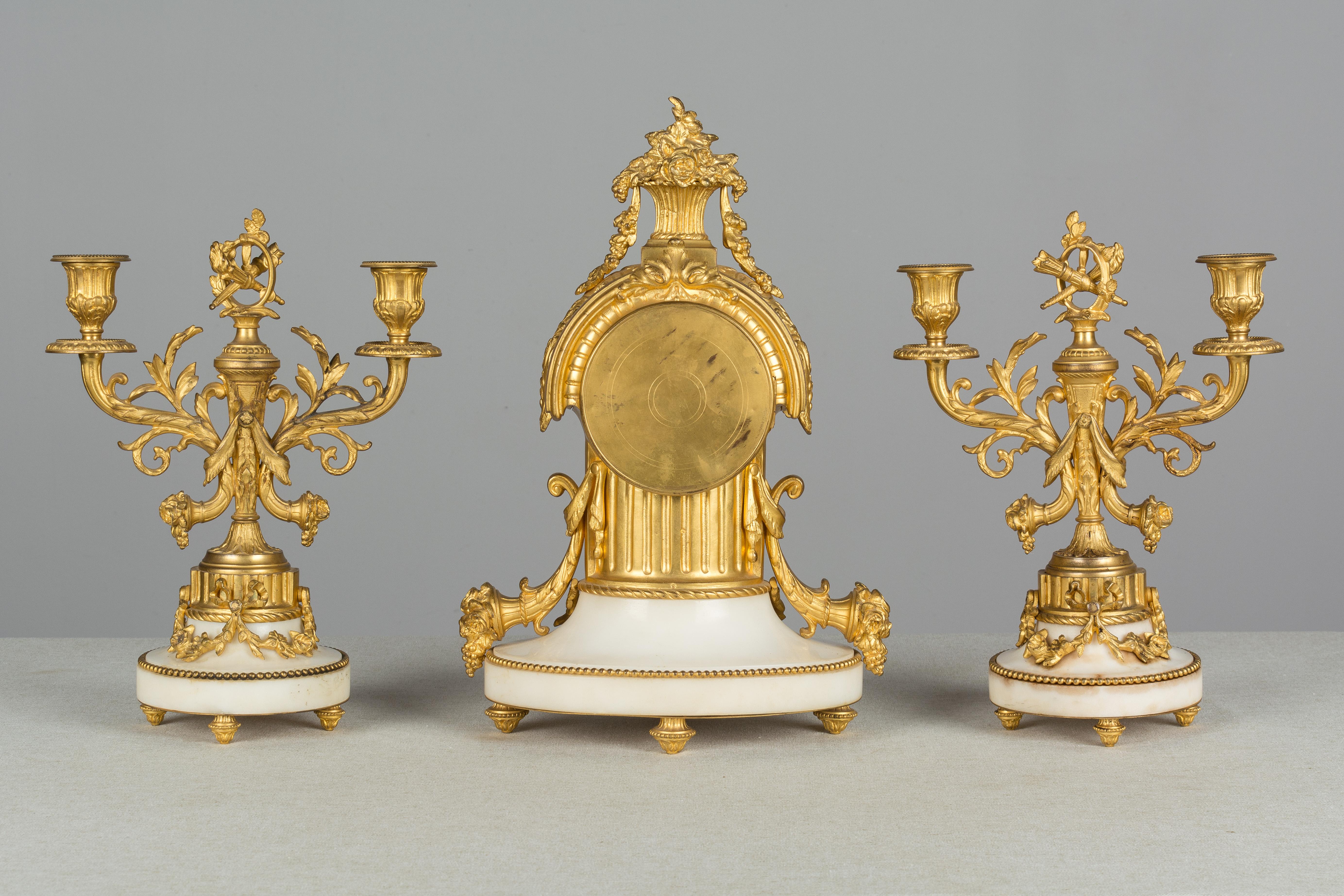 A beautiful 19th century Louis XVI style three-piece mantel garniture consisting of a clock and a pair of two-light candelabras. Bronze doré with Fine casting and bright gilt with white marble bases. Clock has an enamel face, hand-painted with Roman