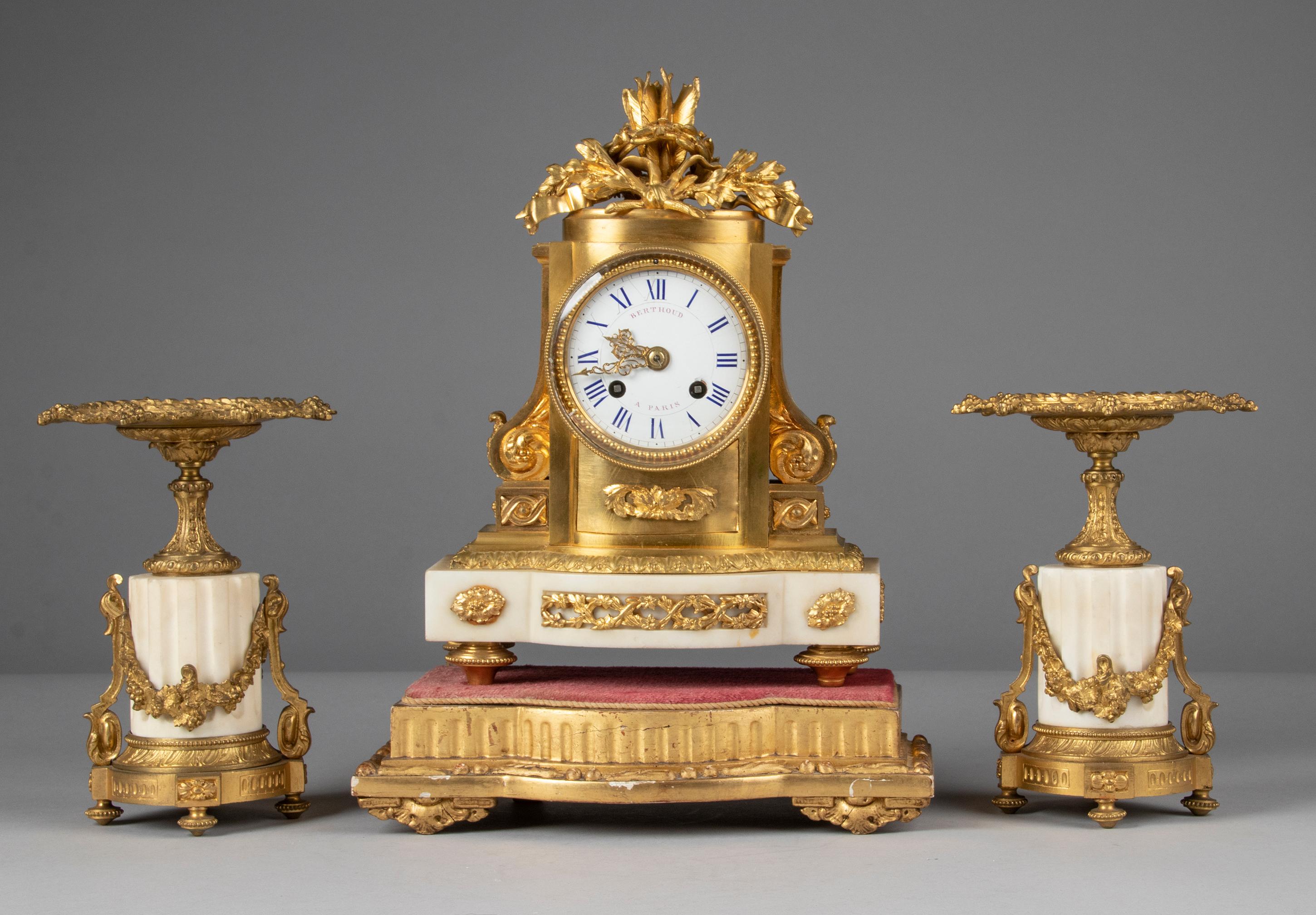 A refined Louis XVI style mantel clock with matching candelabras. The case is made of gilt brass and bronze ormolu casted ornaments, the dial is enameled iron. Resting on a white marble base. Richly embellished with neoclassical ornaments, flower