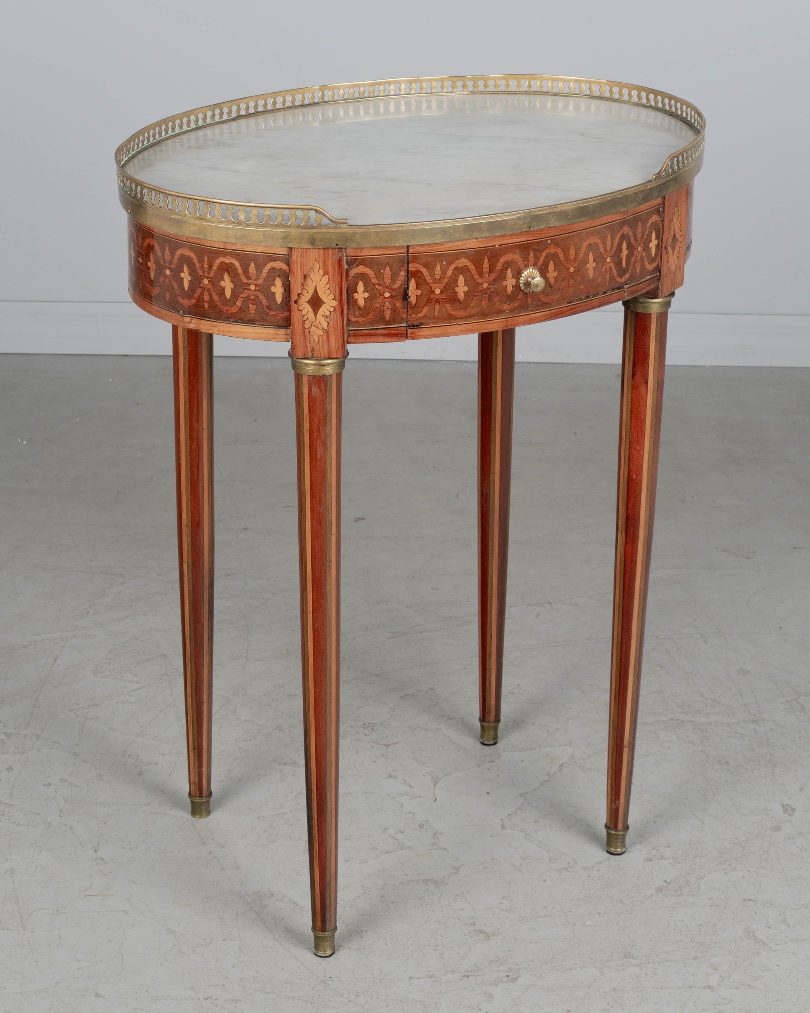 Tavolino ovale con piano in marmo intarsiato in stile Luigi XVI del XIX secolo, realizzato con impiallacciature di mogano, noce e vari legni esotici. Piccolo cassetto a coda di rondine con pomello in ottone fuso. Piano in marmo bianco con galleria