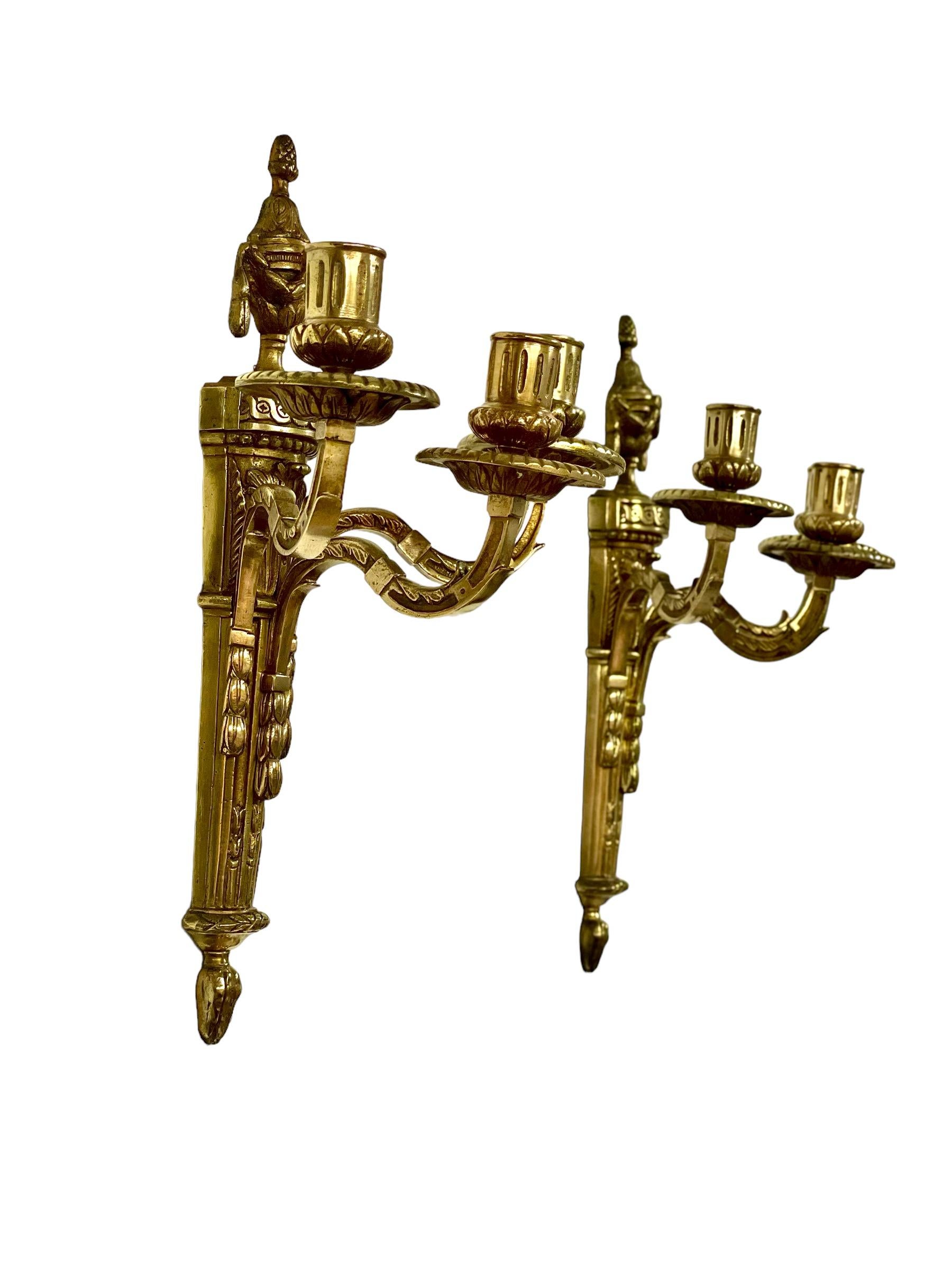 A pair of 19th century, Louis XVI style wall sconces, in chiselled and gilded bronze, in a very striking neoclassical design. The reeded backplate is in torch form, with a swagged urn above, topped with an acorn finial. The two arms have foliate