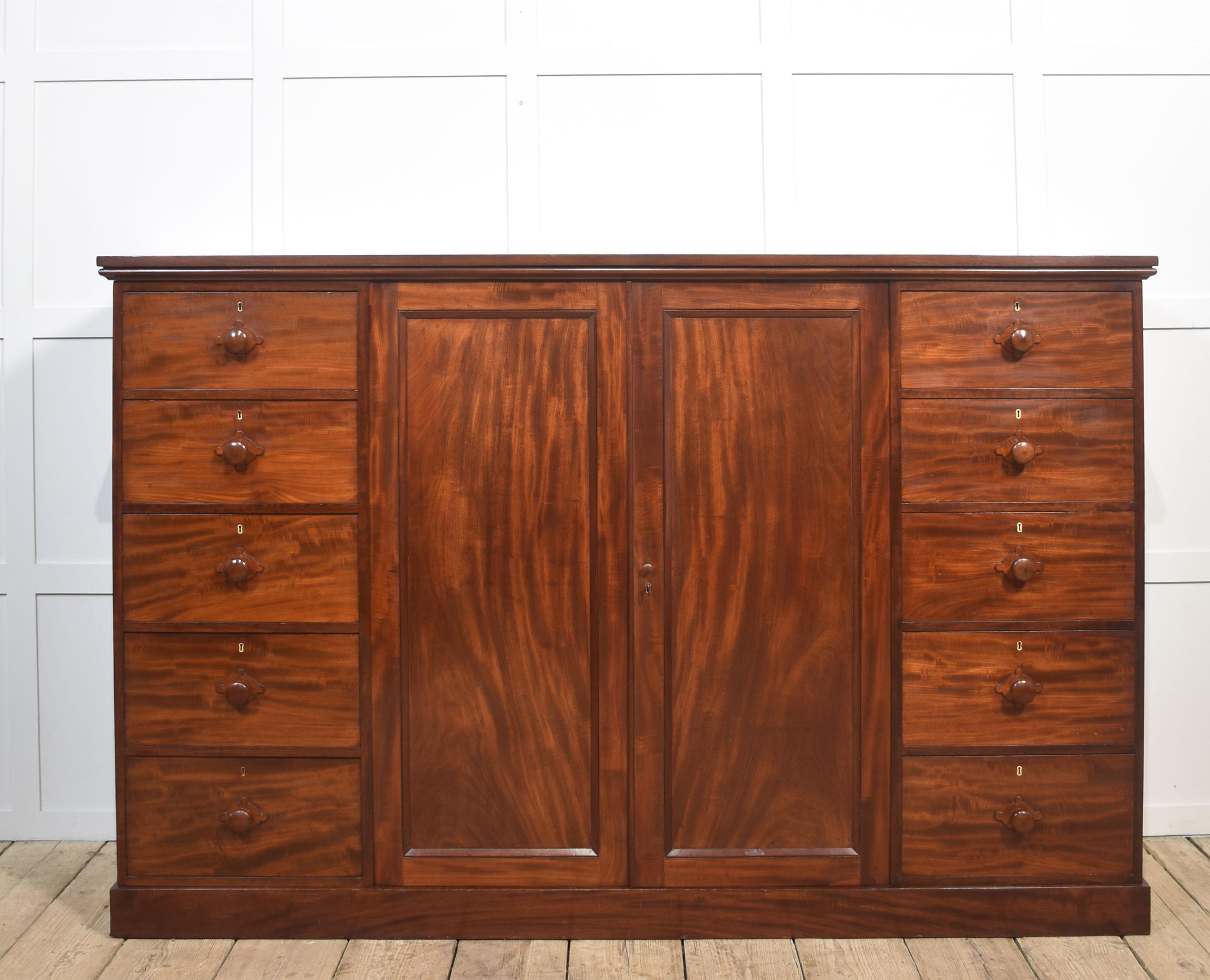 Armario prensero enano de caoba de la Regencia, con una gran tapa rectangular moldeada sobre dos puertas centrales de paneles de caoba flameada. Se abre para revelar un interior transformado con tapicería de ante y una larga barra para colgar.