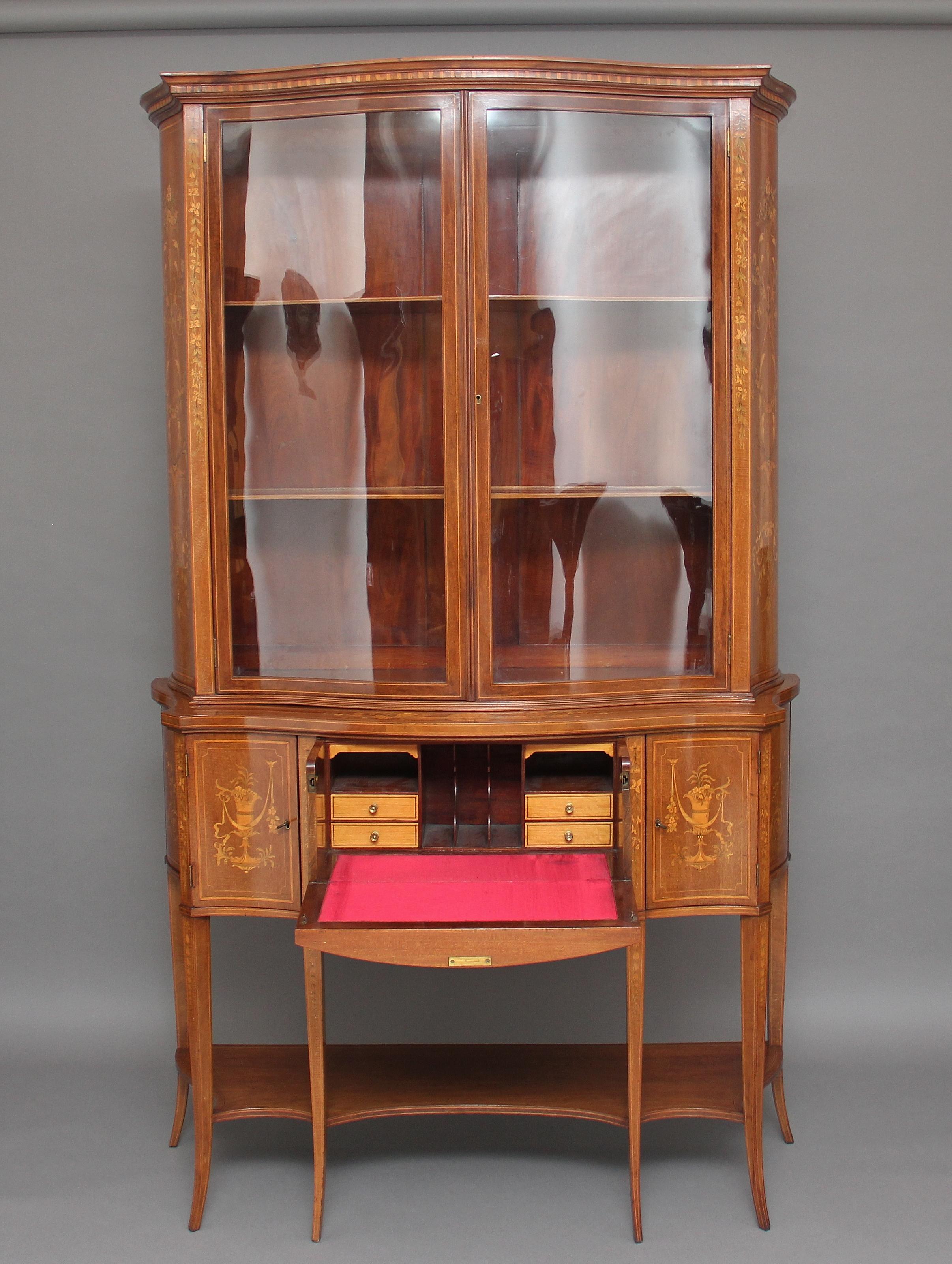 A large 19th century mahogany Sheraton Revival inlaid display cabinet of serpentine form, the shaped inlaid cornice above two serpentine shaped glazed doors opening to two fixed shelves inside, the sides of the top cabinet profusely decorated with
