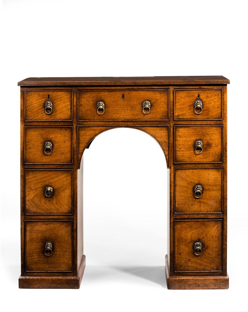 A charming kneehole desk, possibly made for a young man. Comprising of nine oak lined cock-beaded drawers, each with brass lion mask ring handles. The whole is raised on a small plinth base. This article will make an ideal side or lamp table,