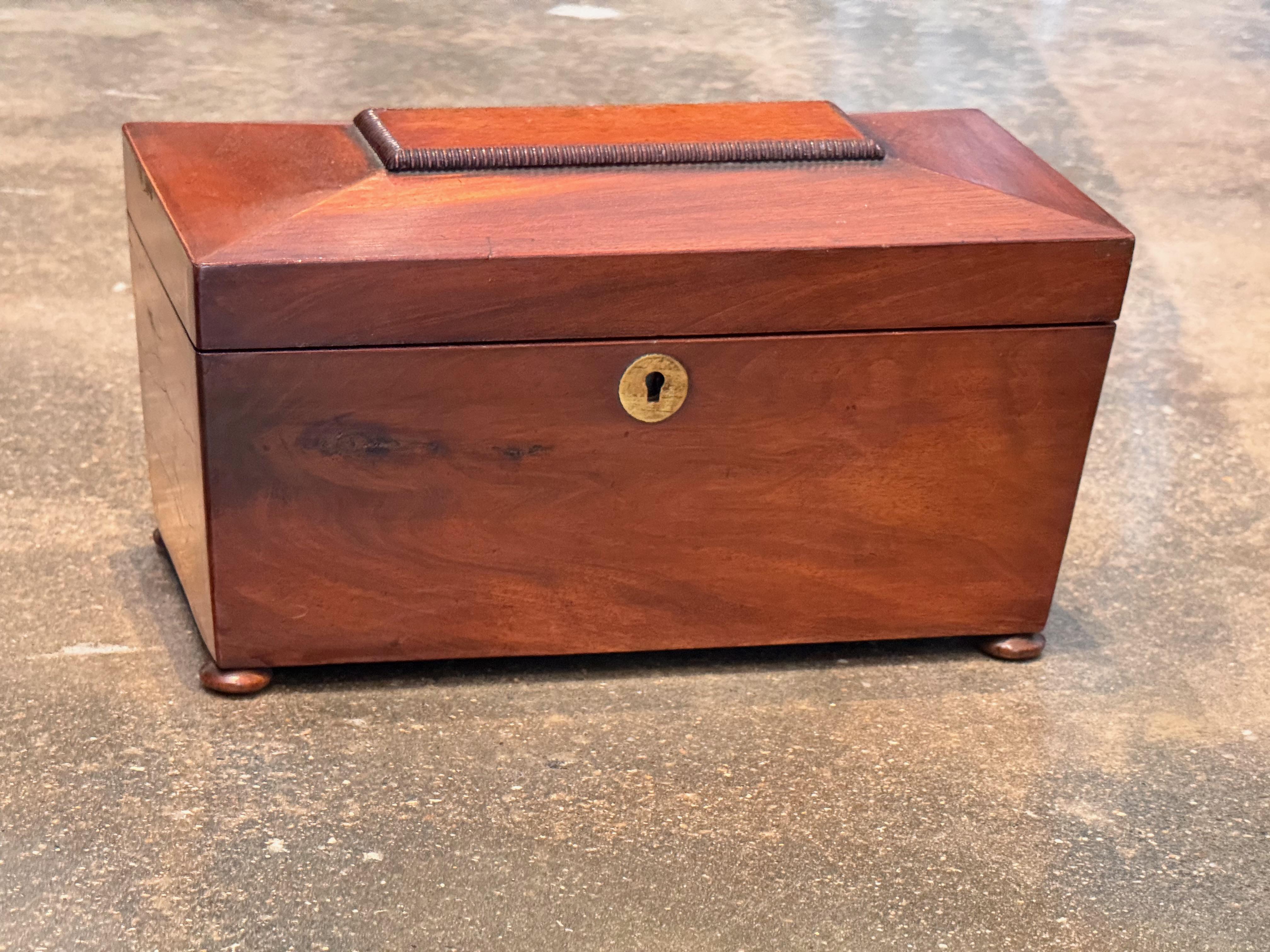 Rectangular mahogany tea caddy featuring a beveled lid with raised handle, resting on small bun feet. The interior is lined in red and fitted with two lidded compartments for storing loose tea. The box retains its original lock hardware, and the