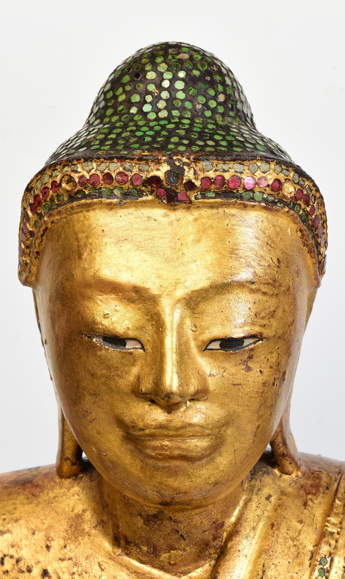 Bouddha ancien en bois de Birmanie assis en posture Mara Vijaya (appelant la terre à témoin) sur une base laquée, dorée et incrustée de pièces de verre colorées sur la tête, le bandeau et les bordures de la robe. 

Âge : Birmanie, période Mandalay,