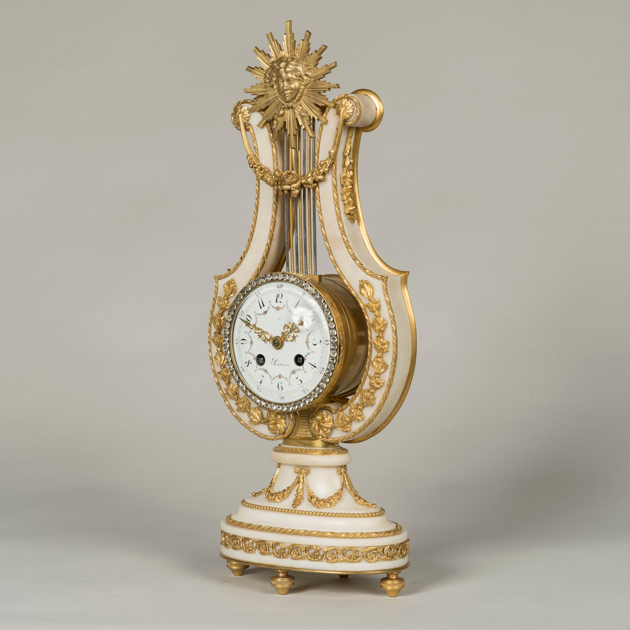 A French Clock Garniture
in the Louis XVI Manner

The clock and its accompanying candelabra constructed in gilt bronze mounted Italian Carrara marble; the timepiece with stepped elliptical base rising from toupie feet, dressed with running rinceaux