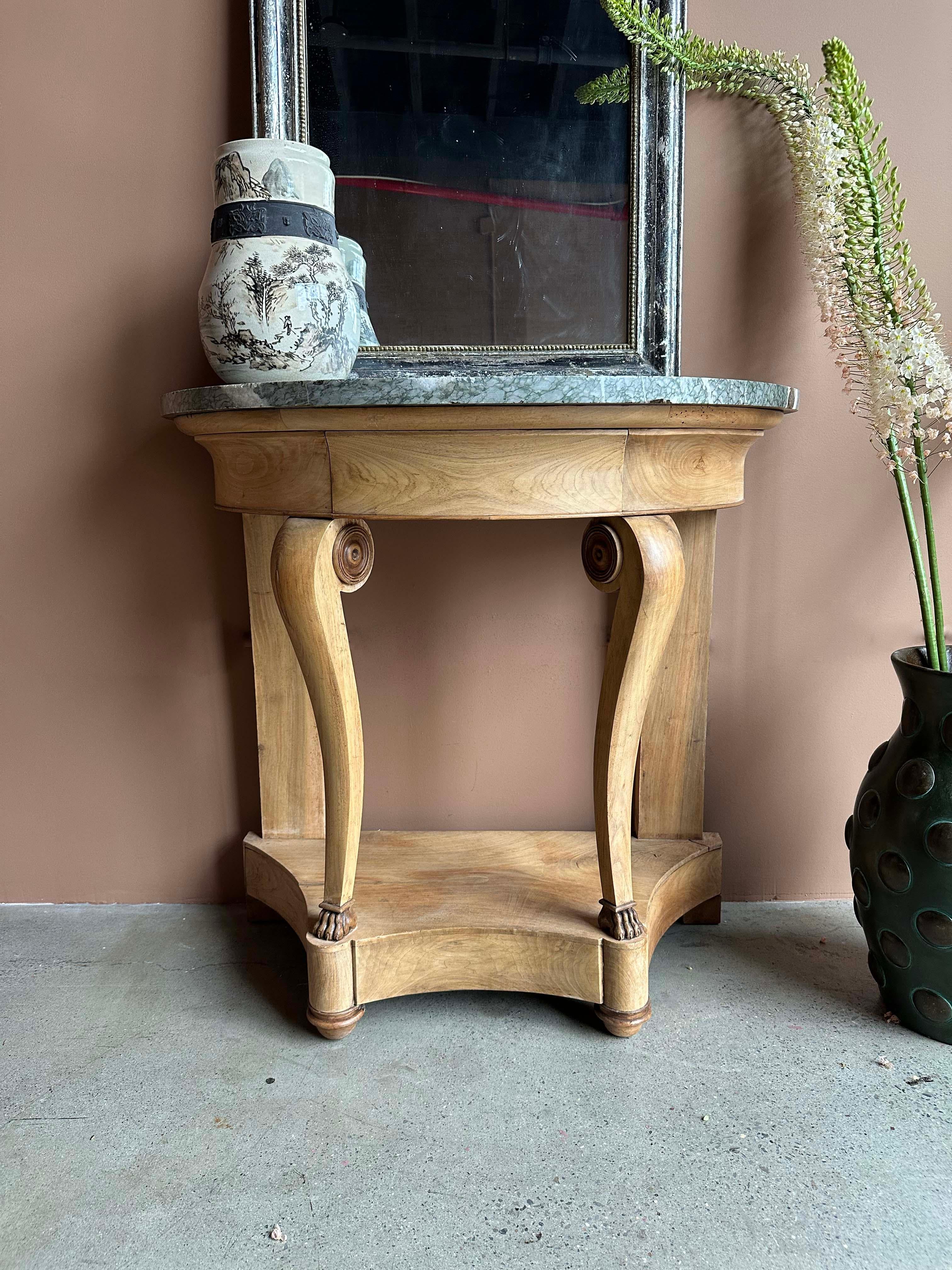 An elegant 19th-century demi-lune console table featuring a richly veined marble top over a carved blond wood base. The neoclassical form is distinguished by scroll-accented front legs terminating in paw feet and joined by a shaped lower shelf, all