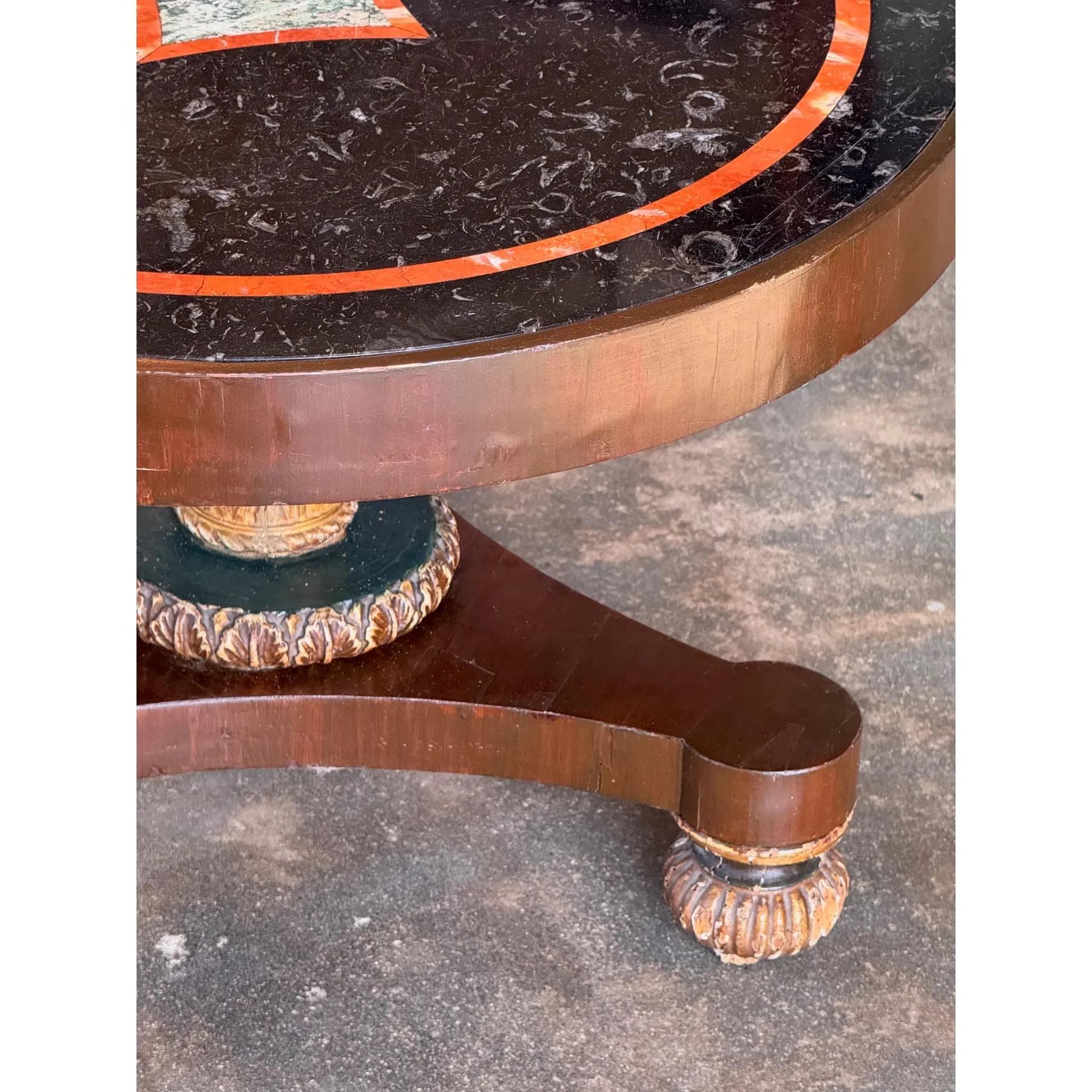Round gueridon table featuring a striking black marble top with an inlaid border and central decorative panel. The rich wood base is supported by a pedestal with a painted and gilded carved foliate design, leading to a tripod base with carved feet.