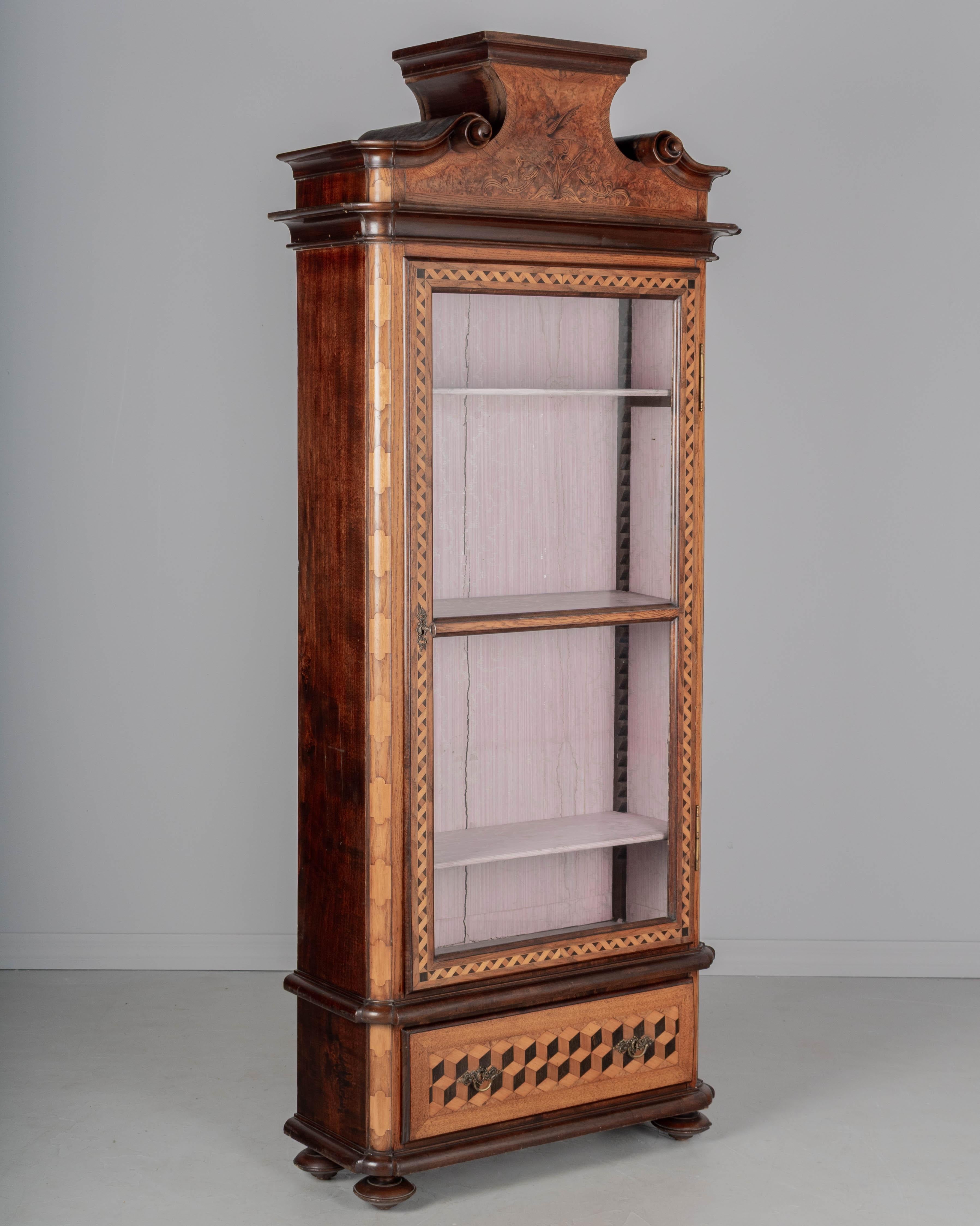 Vetrina o espositore in intarsio del XIX secolo proveniente dalla Svizzera. Realizzato in noce con impiallacciature intarsiate di acero e olmo. Grande corona scultorea con volute intagliate a mano e uccello intarsiato. Porta a vetri con serratura e
