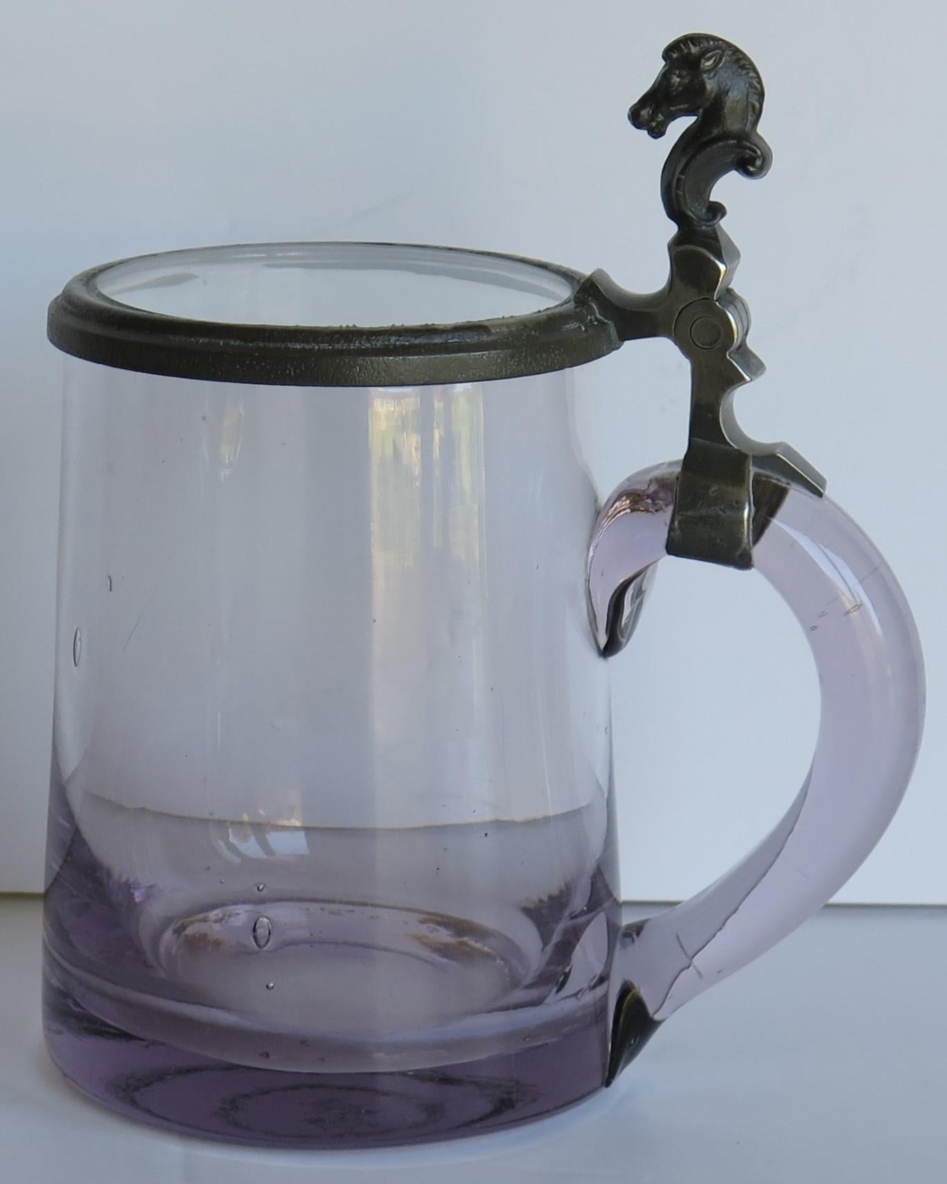 This is a good quality hand blown glass tankard with a hinged cover or lid having a pewter horse head thumb piece, crafted in the mid-19th Century.

The tankard has a cylindrical slightly tapered body which is made of hand blown glass with a good