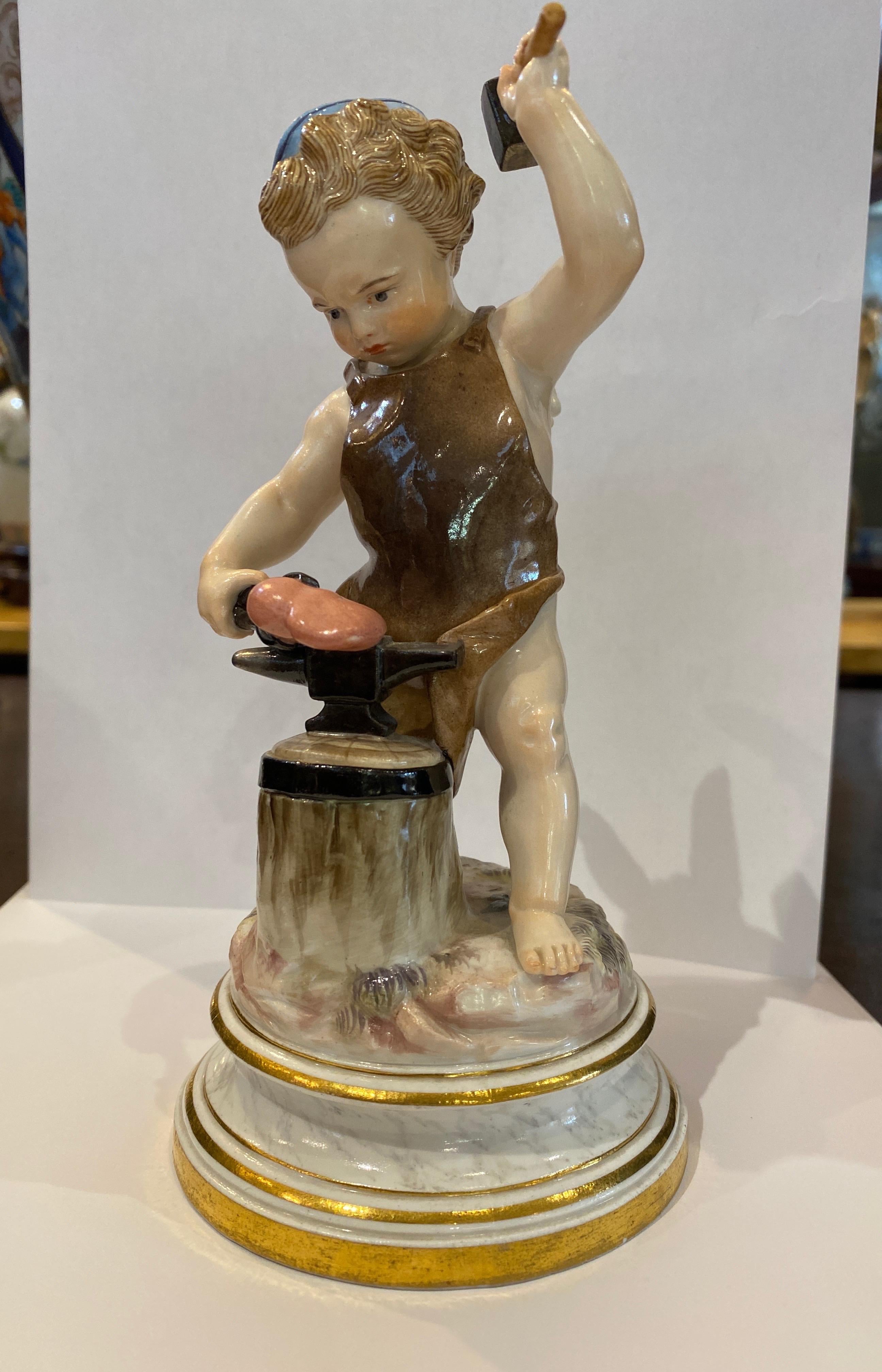 Meissen Porcelain figure of cupid forging a heart on a blacksmiths anvil which rests on a block of tree stump. The barefooted figure wearing a dark blue cap and dressed in a brown leather smock, mounted on a circular marbled base with gilt
