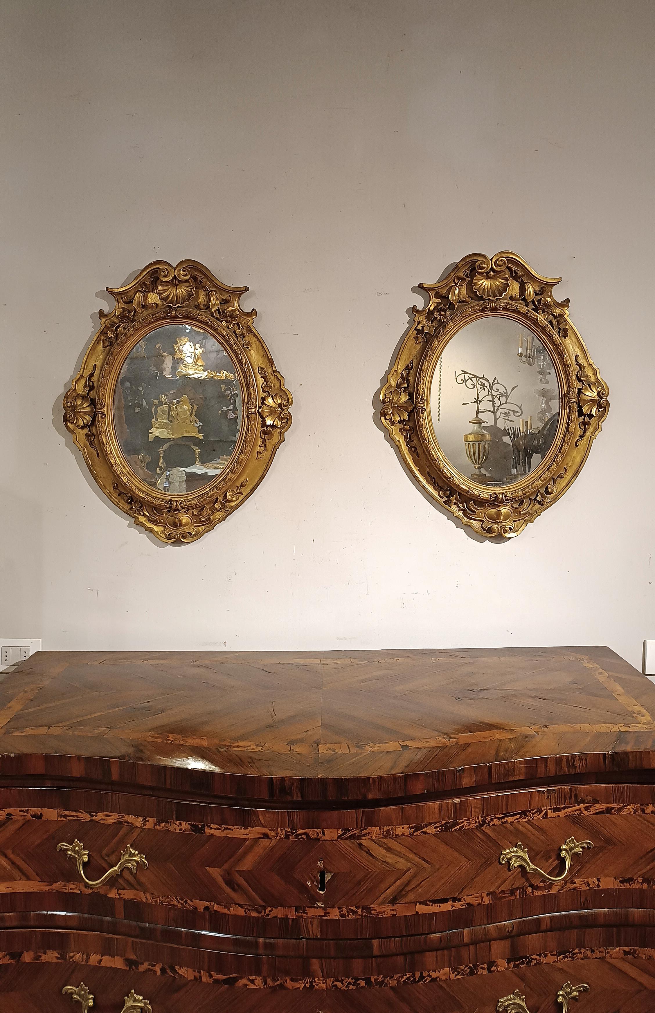Beautiful and elegant pair of oval mirrors in carved and gilded pine wood with pure gold leaf, which adds a touch of opulence and refinement to them. The decoration includes shells and phytomorphic elements, creating a fascinating and unique design.