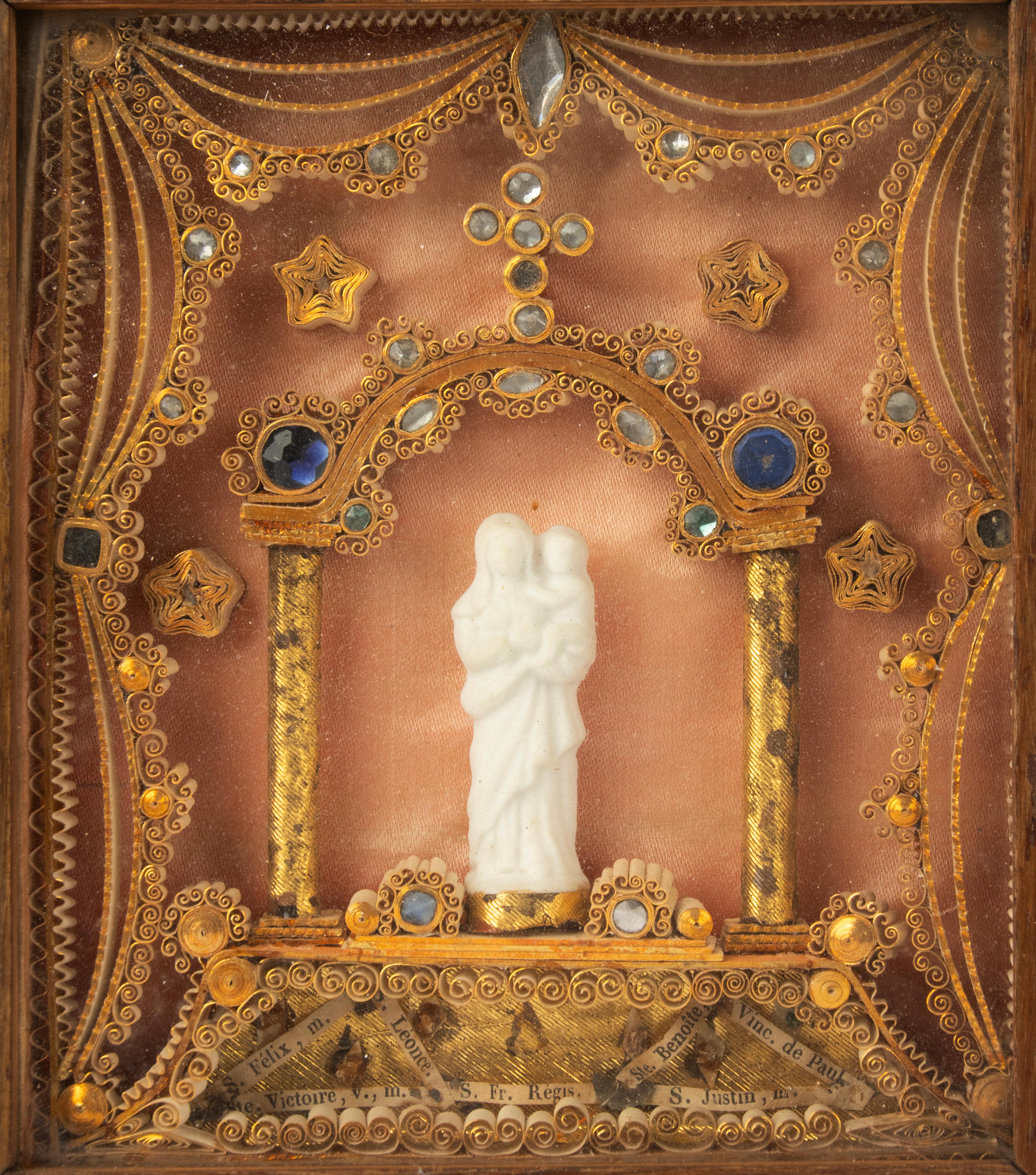 Cadre reliquaire de dévotion du XIXe siècle, français, contenant plusieurs reliques sous verre. Au centre se trouve une délicate figure de la Vierge à l'Enfant en porcelaine blanche, encadrée par deux colonnes dorées et un dais arqué richement