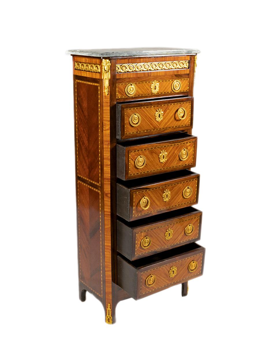 Chiffoniera a cinque cassetti in stile transitorio del XIX secolo, con piano in marmo e montature in bronzo dorato, maniglie e marqueterie della cassettiera a sbalzo con angoli in bronzo, fianchi a pannelli con parquet abbinato. 
Si regge su gambe