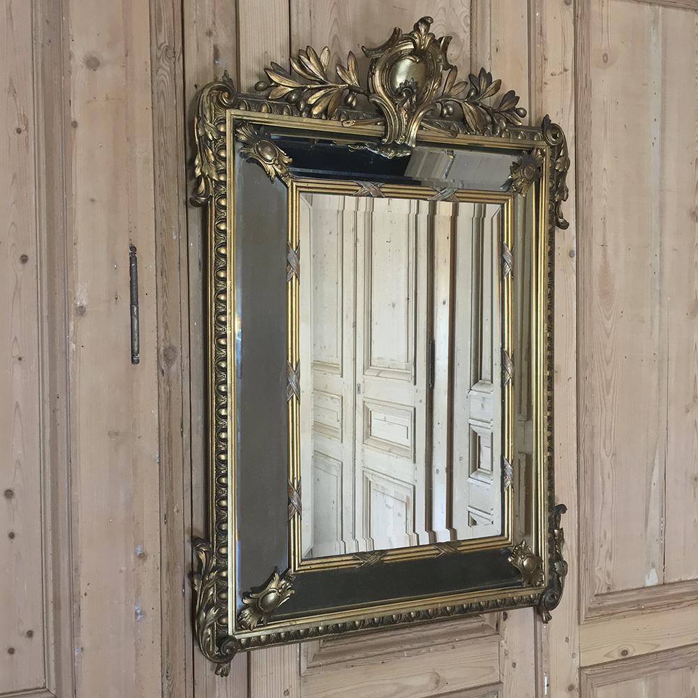 Impeccable 19th century antique neoclassical French gilded mirror boasts a crest motif as a cartouche centered on the top, with olive branches emanating from the crest. Handsome acanthus plumes adorn the shoulders and grace the corners of the base,