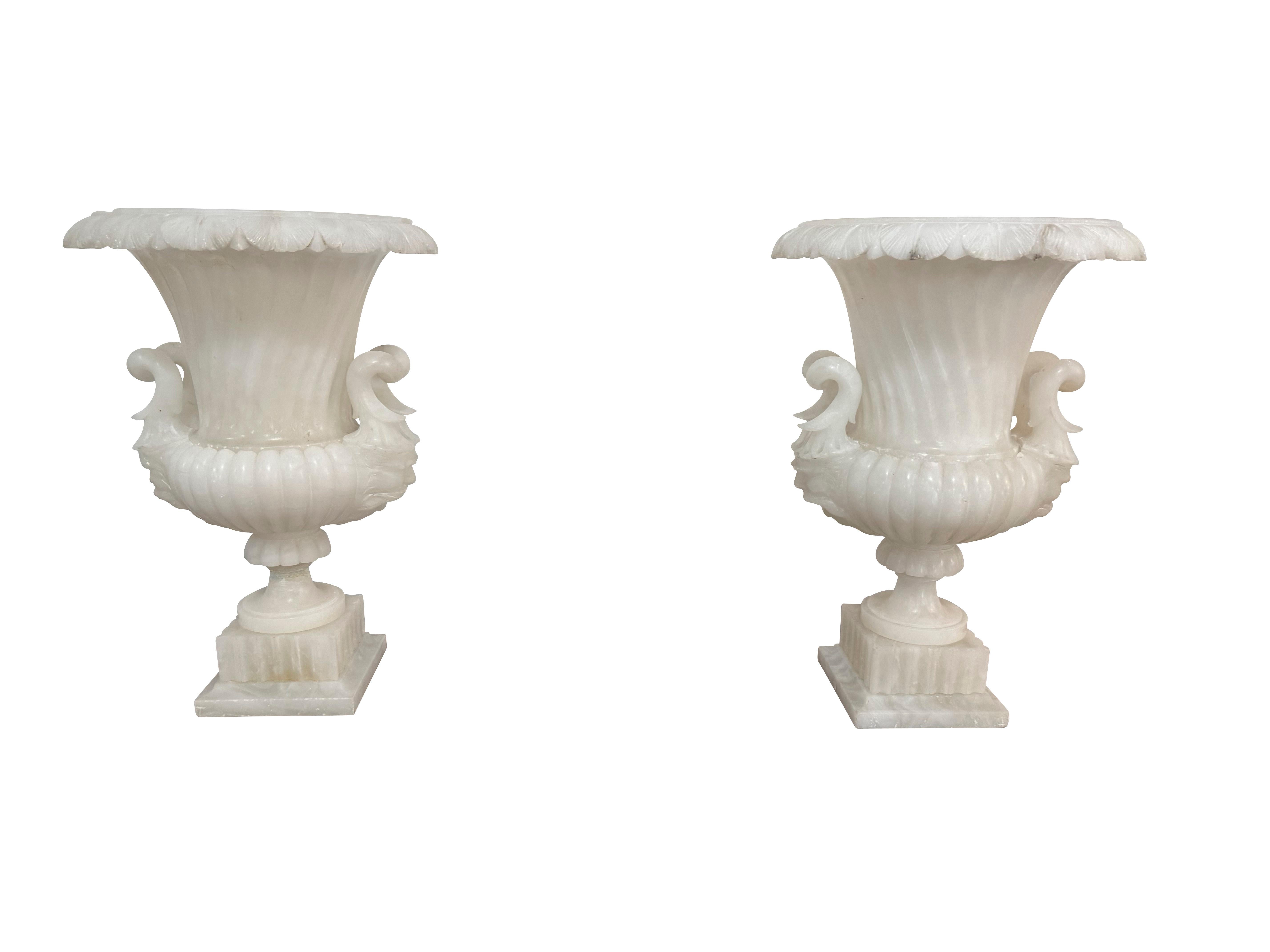 A Unique pair of Neoclassical hand-carved Alabaster urns in two pieces with pedestal base and urn form top.  The graceful outline of the urn is in a h swirl with lovely scroll-form handles on both sides. The lips of the urns are heavily carved with
