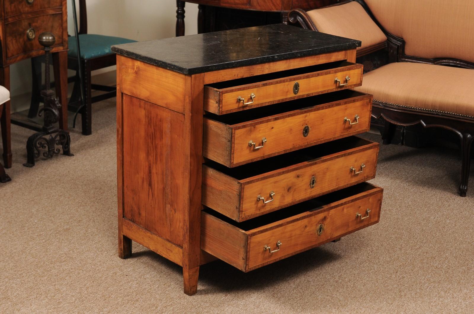 The neoclassical style petite Italian fruitwood commode with black marble top, 4 drawers with inlay/cross-banding and brass pulls terminating in tapered feet.

