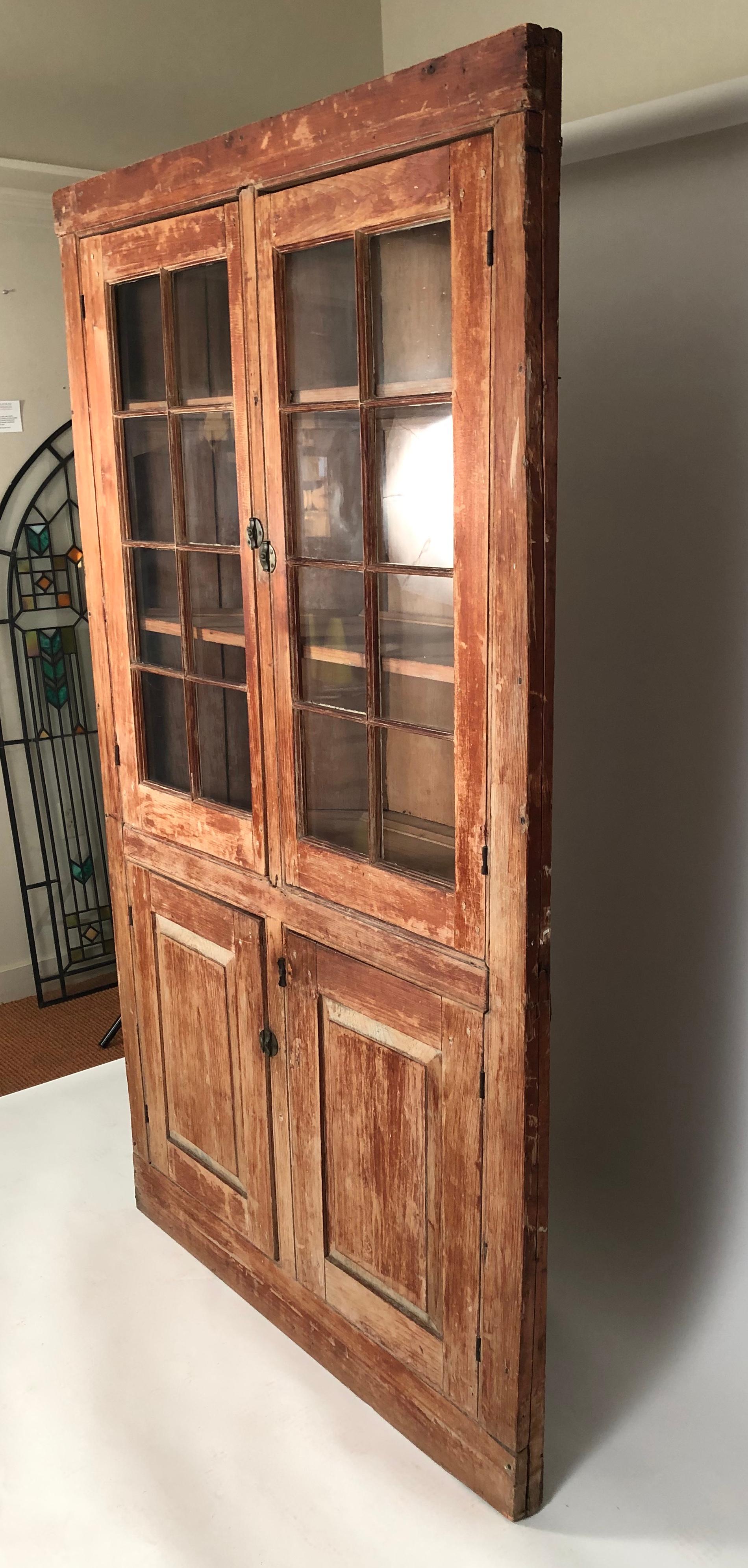 A 19th century American painted country corner cupboard, circa 1840, with wonderful old surface, in pine with layers of original old red and blue paint, in one piece, the upper section with two glazed doors enclosing three shelves, each with a plate