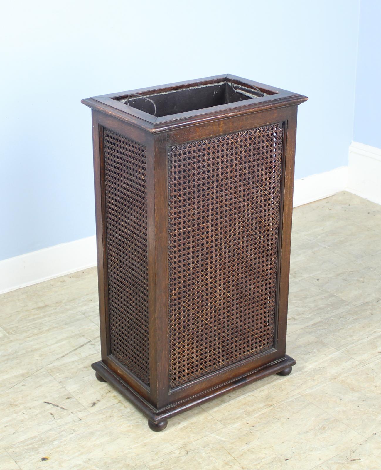 A good simple and compact stick or umbrella stand on four bun feet with moulded platform base. Caned sides and front enclosed in oak frame. Original metal liner with original handles.