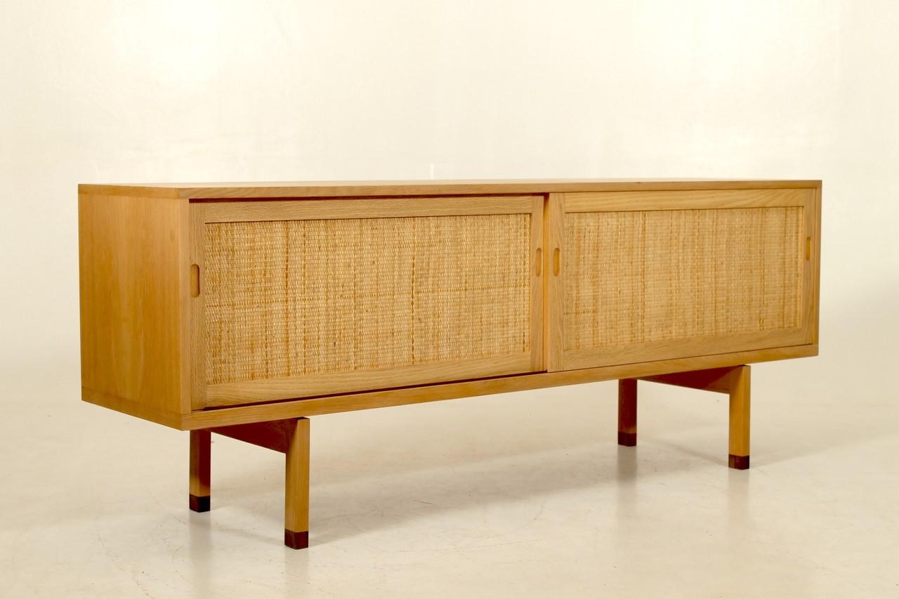 Low Hans Jørgen Wegner sideboard with oak veneer front with two solid sliding doors with woven cane panels, enclosing shelves and trays, solid oak legs with dark wood leg ends. Produced by Ry Møbler, 1960s.You won't find this special old low