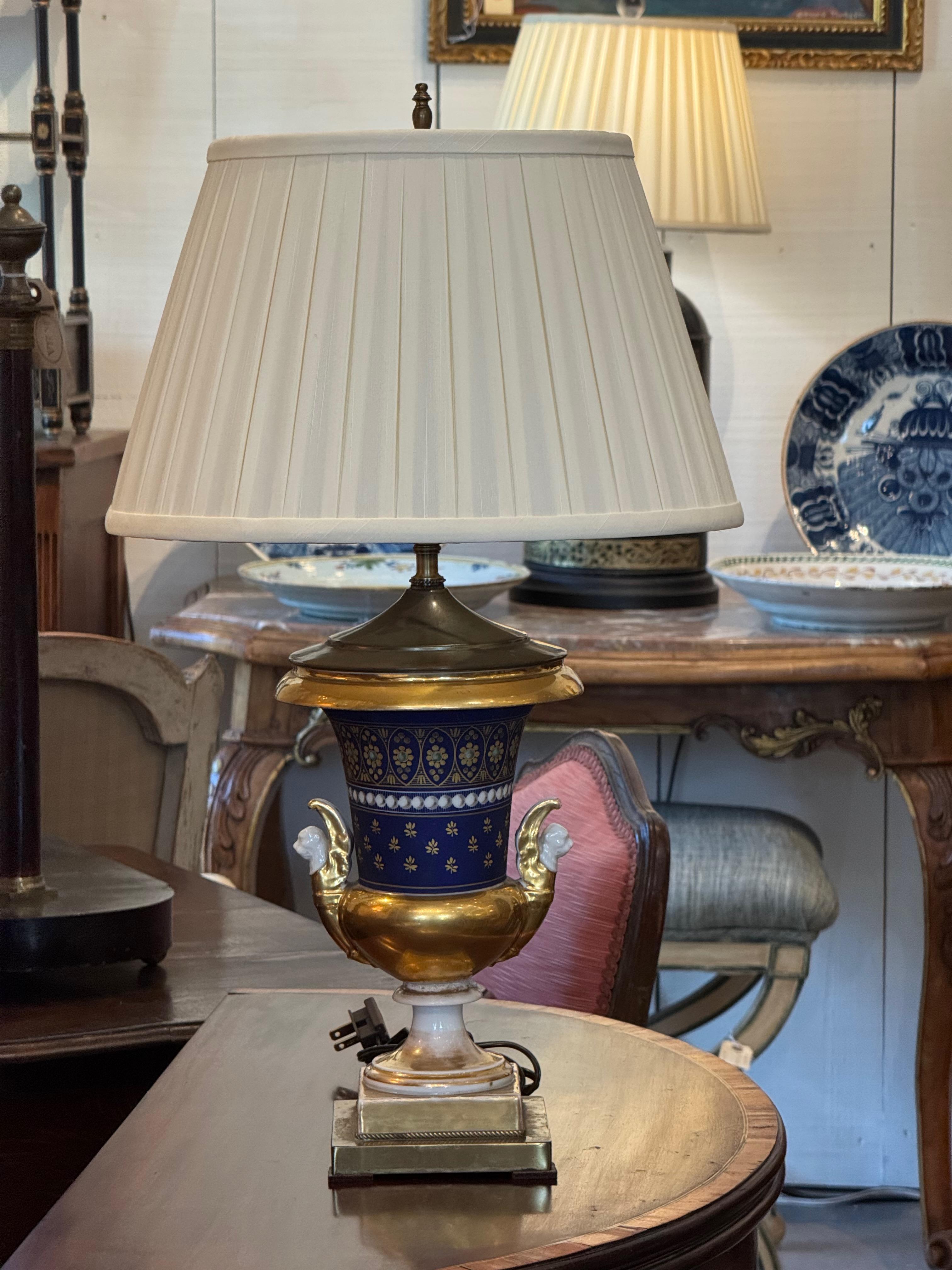 Lámpara urna de porcelana Old Paris con cuerpo azul cobalto, decoración clásica dorada y asas blancas en forma de cisne. Montada sobre una base cuadrada de estilo neoclásico y equipada con una pantalla plisada de color crema, esta lámpara aporta la