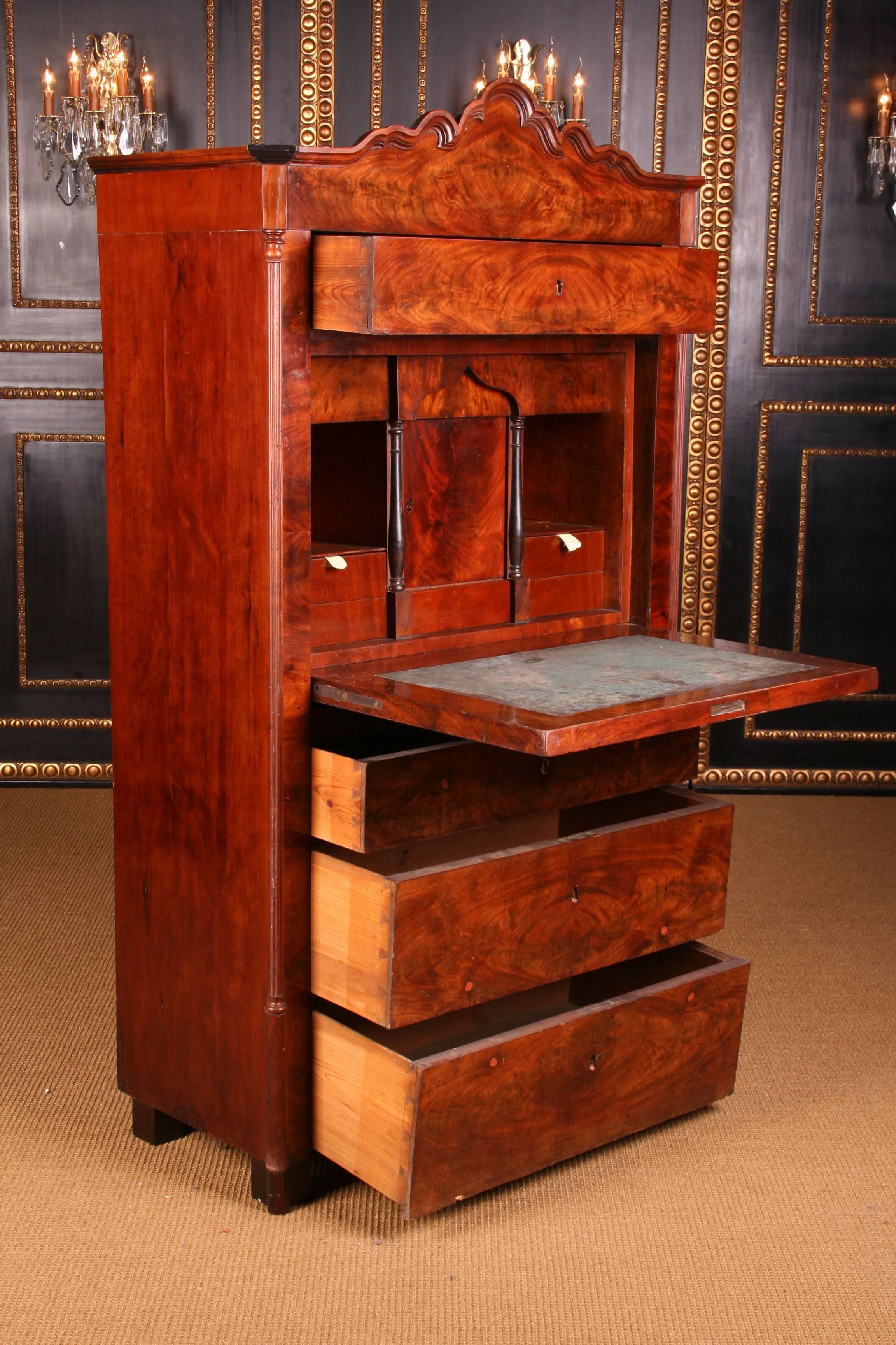 Mahogany on solid pinewood. High-right body on block-feet. Straight writing plate with a medallion-shaped depression. Behind it, architecturally designed interior. Curved cornice, flanked by Amphora-shaped ornamental elements. This secretaire is in