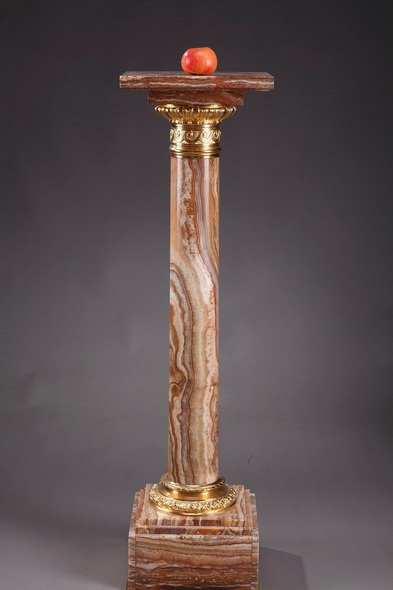 Onyx display column in a spectrum of ochre, orange and yellow tones. The top and bottom of the shaft is encircled with a ring of gilt bronze that is embellished with alternating palmettes and ova on the bottom and a frieze of waves on the top. The