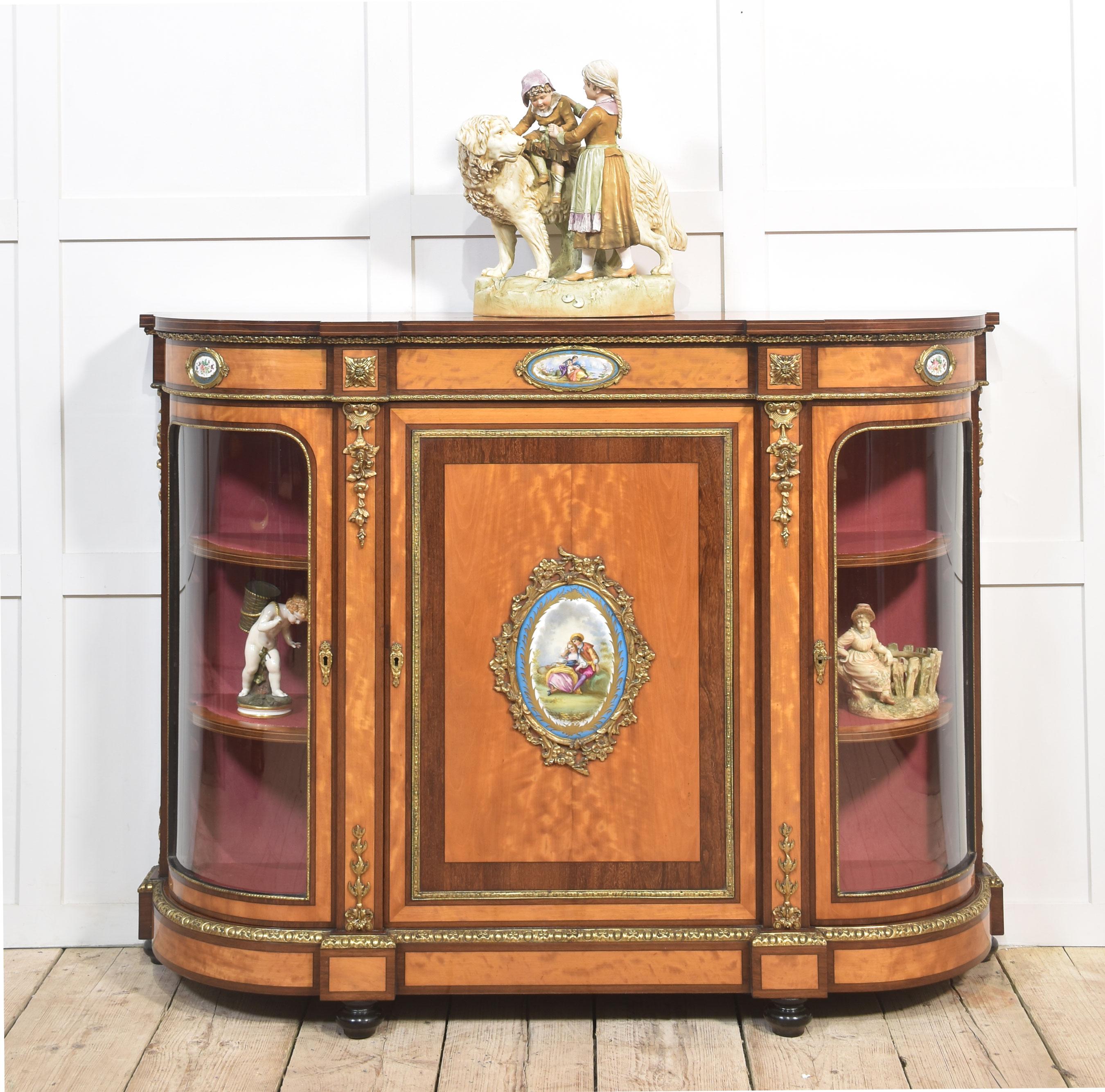 Aparador de madera de satén, montado en ormolu, con la parte superior bien labrada y el borde reticulado. Sobre una puerta central con panel ovalado de porcelana de Sevres. Los extremos están equipados con puertas acristaladas arqueadas que