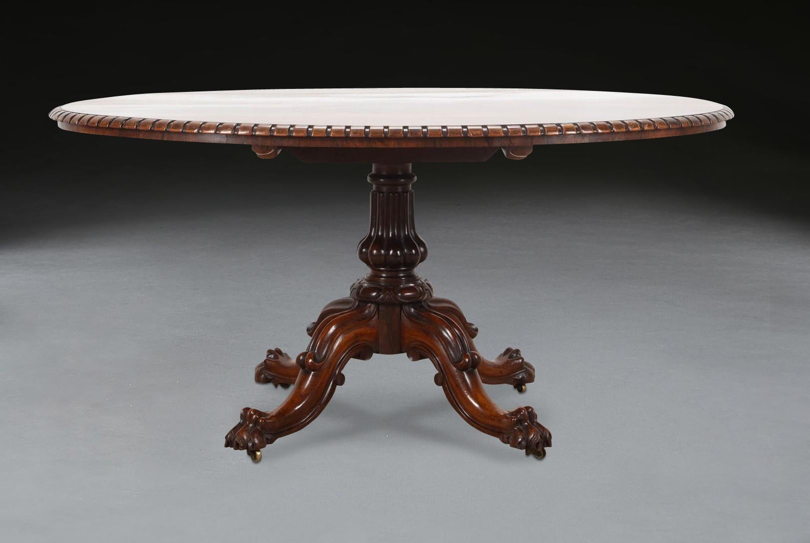 Table centrale ovale en palissandre du XIXe siècle, de belle forme, le plateau basculant à bord godronné et à frise peu profonde, reposant sur une colonne sculptée en forme de vase à quatre pieds évasés, avec des lappets sculptés sur les genoux et