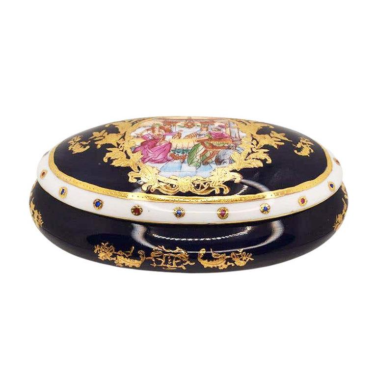 A gorgeous hand-painted oval porceclain box with a highly decorated lid. This lovely Neoclassical Revival piece is created from porcelain, and features an oval shape, with a removable lid. The lid depicts a romantic scene of the French aristocracy