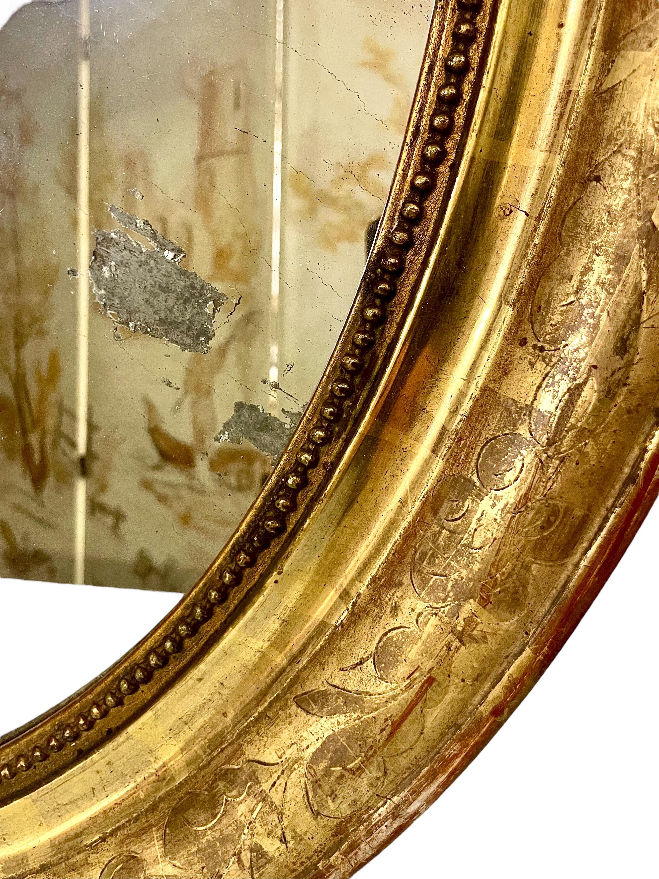 Miroir mural ovale d'époque Napoléon III, dans un cadre doré joliment vieilli, avec une moulure en rang de perles entourant le verre d'origine du miroir. Un élégant motif de fleurs et de feuillages a été finement gravé dans le cadre. De bonne