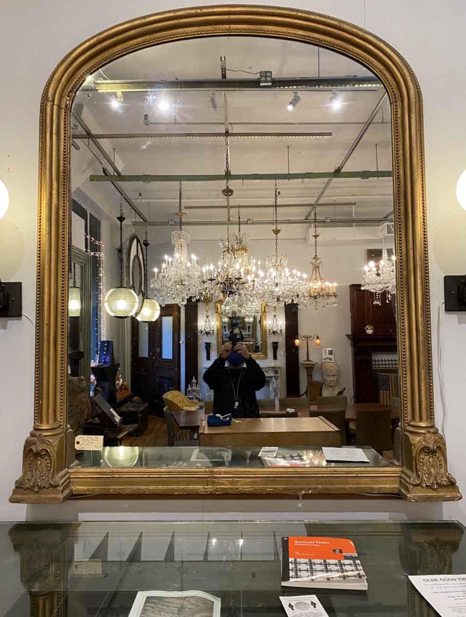 This antique gilded overmantel mirror is from the 19th century. The gilt wood frame has an arched top with two rows of beading running along the edges, which ends in a French design at the extended base. Some minor chipping of the gold leaf.