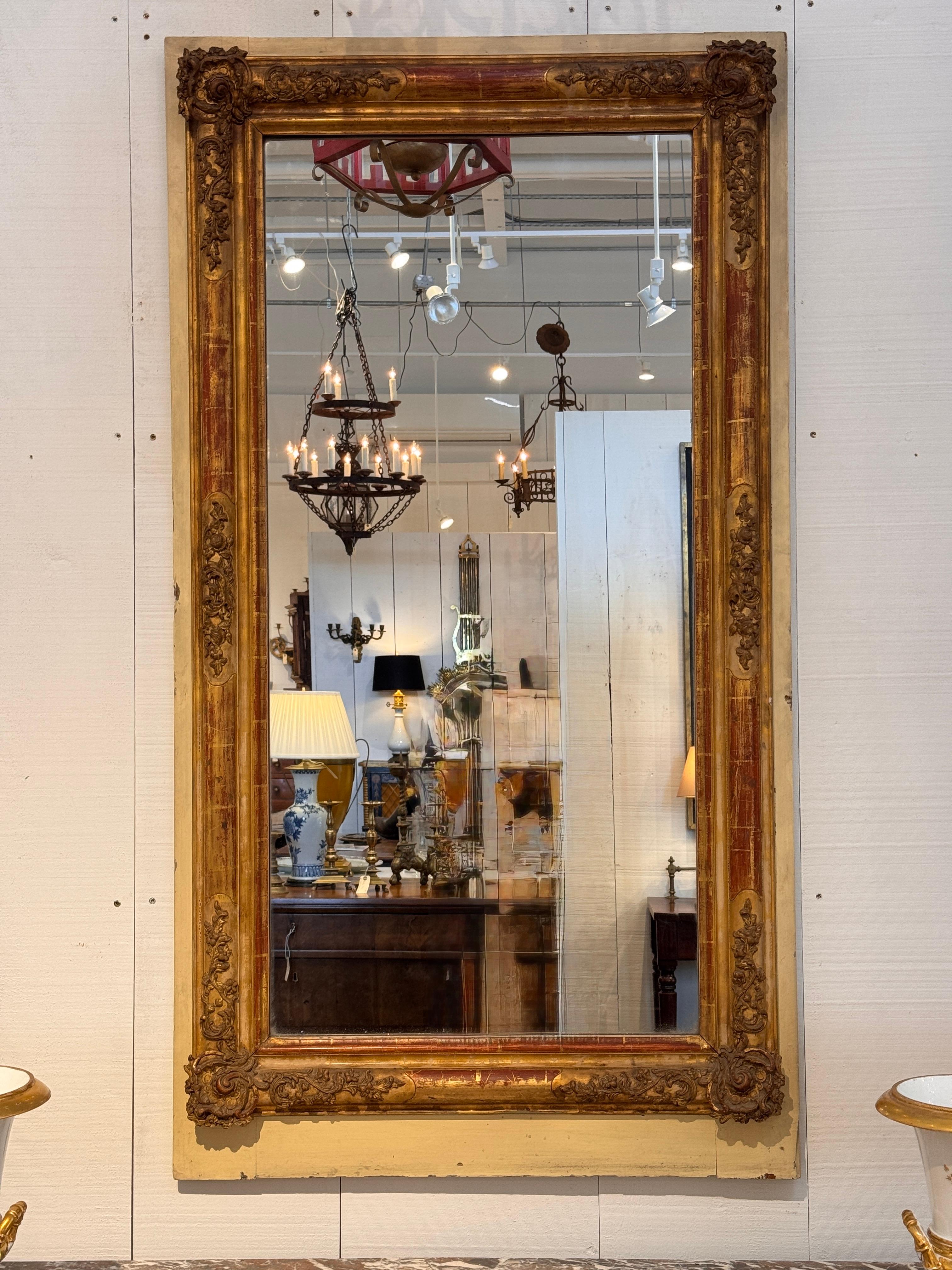 Grand miroir rectangulaire avec un cadre en bois doré richement mouluré, accentué par des sculptures florales et feuillues. La surface présente une patine chaude avec des traces de bole rouge d'origine et de contour peint, ce qui ajoute de la