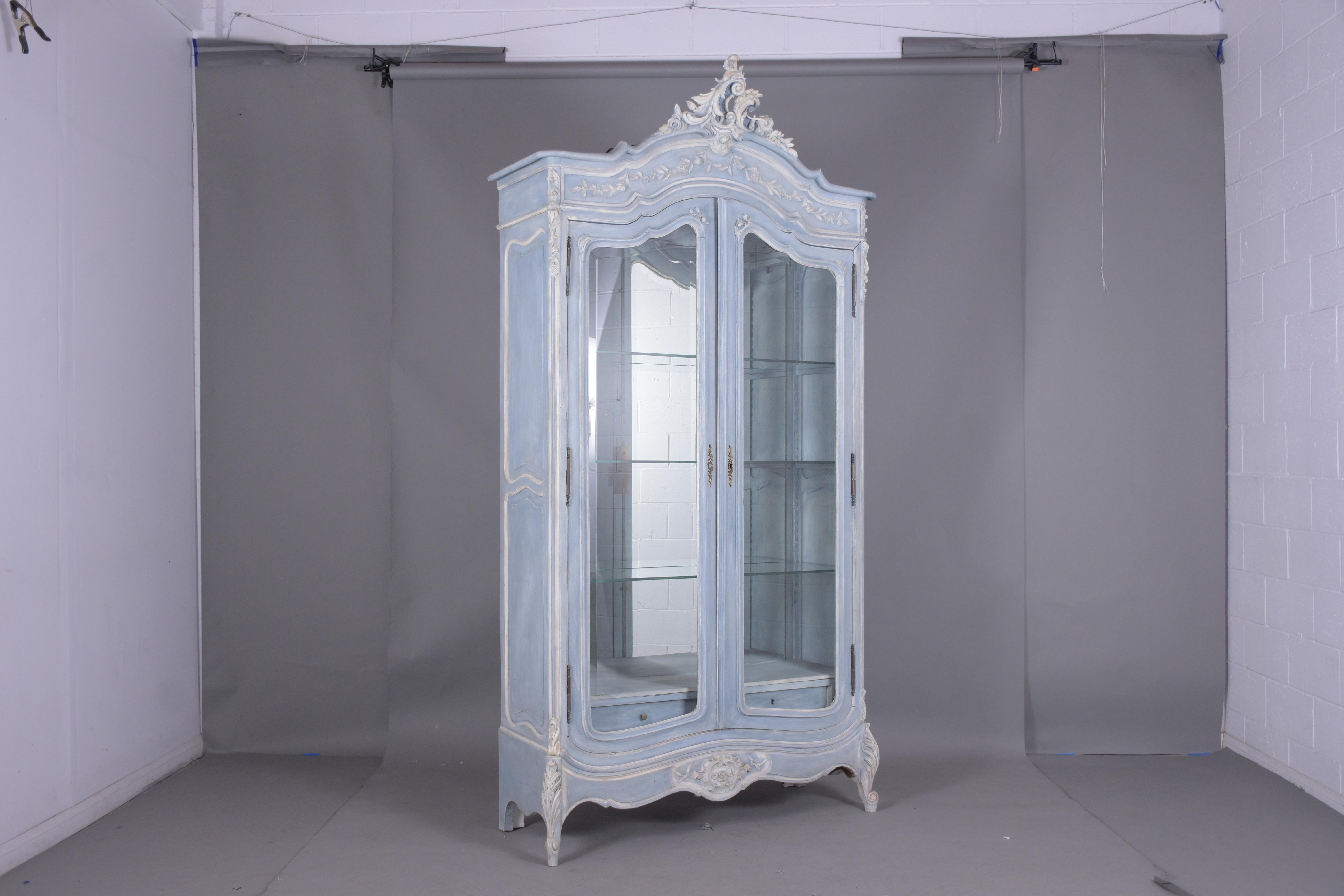 A unique antique French Louis XV display cabinet crafted out of walnut wood with remarkable hand-carved details throughout the entire piece. The bookcase is newly painted in a pale blue and off-white color and features a mirror on the back/bottom