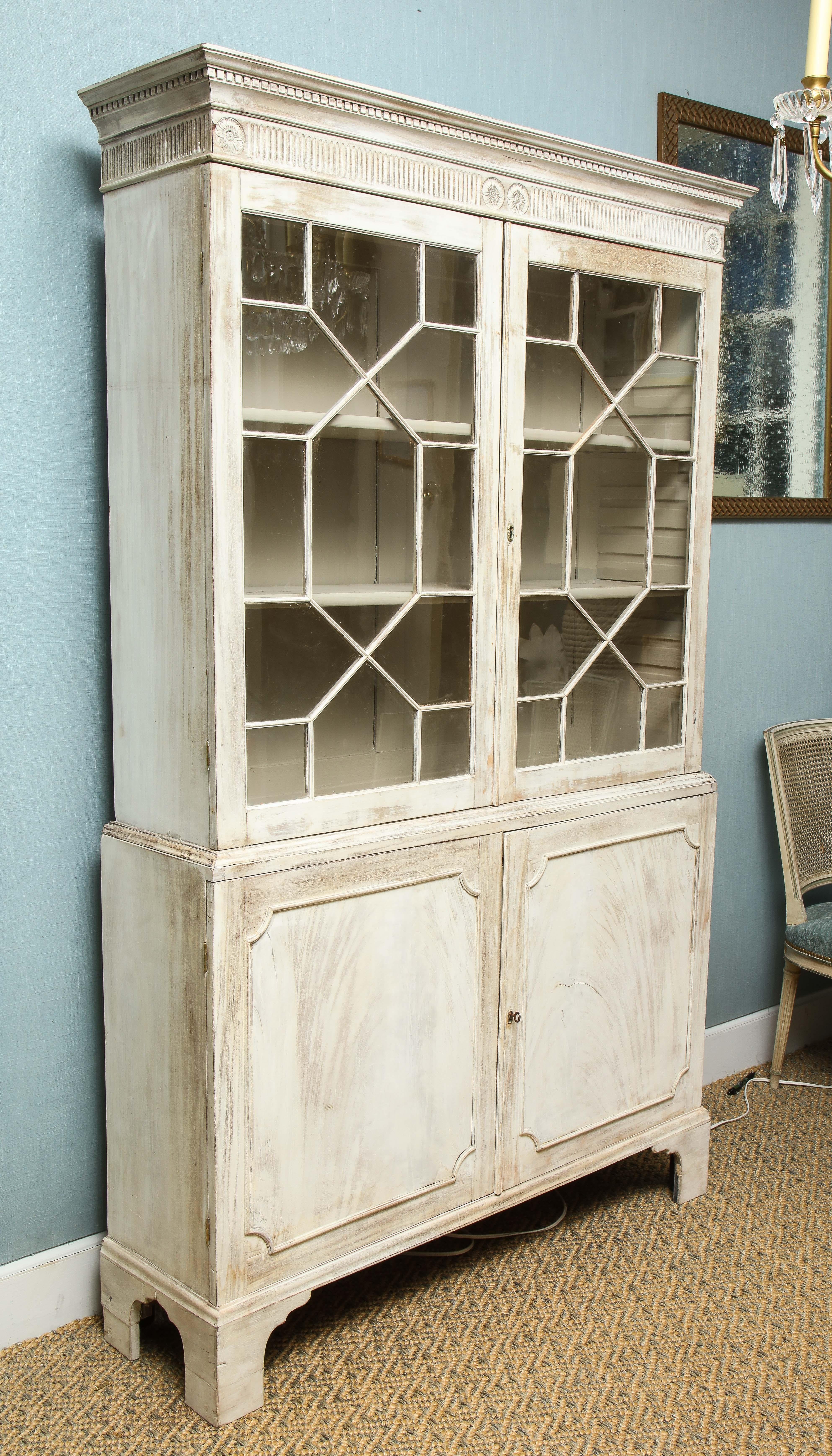 This painted English cabinet has a quiet elegance about it. The piece features two glazed mullioned doors opening to an upper cupboard over two lower paneled doors and two lower storage shelves. Raised on bracket feet, the cabinet features a dental