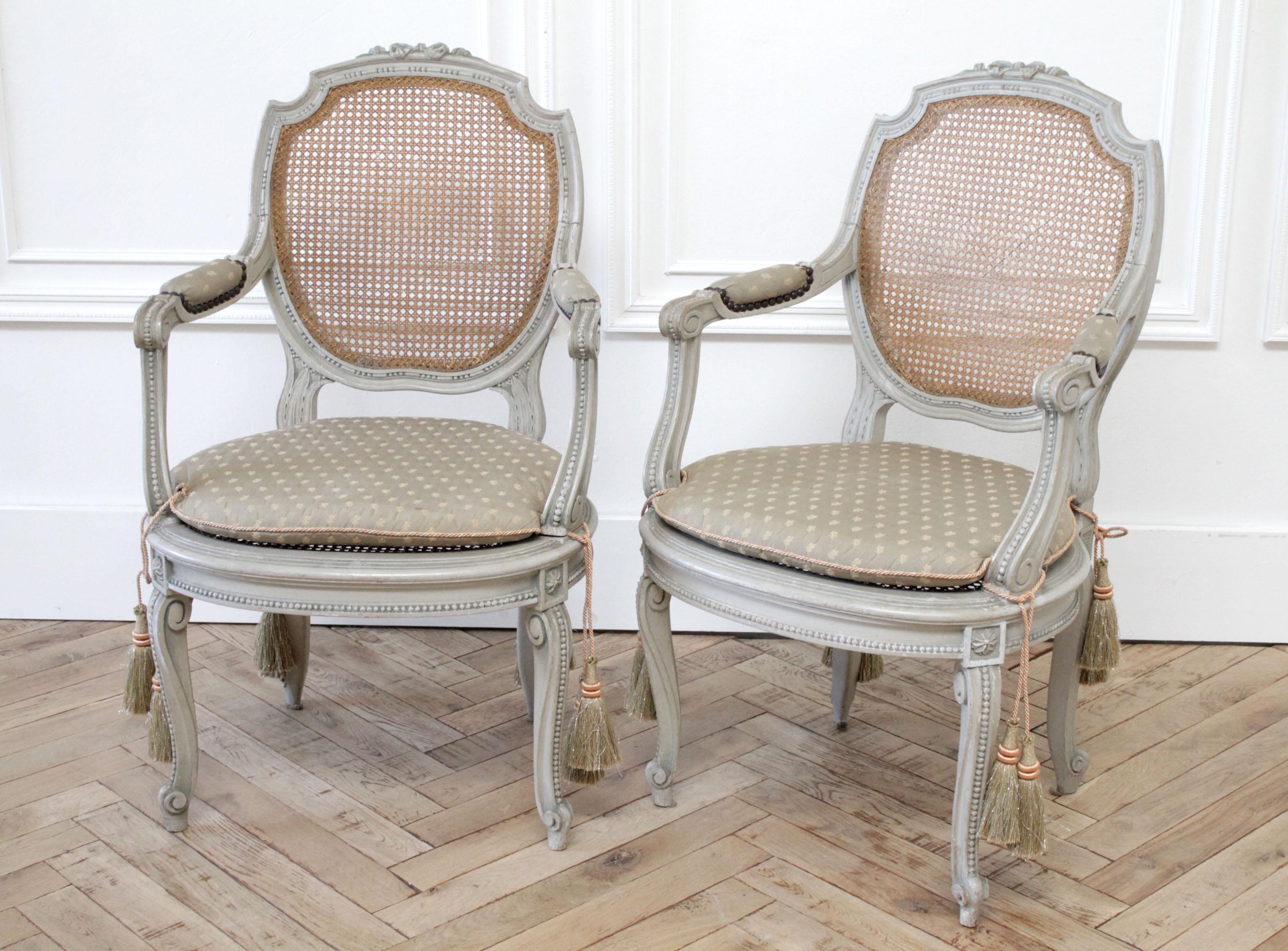19th century painted and carved ribbon cane back open armchairs painted in a soft pale Swedish gray color, with natural cane back and seat. No flaws to the cane back, and seat on one, and the 2nd shows something so minor it’s hard to see. Open arms