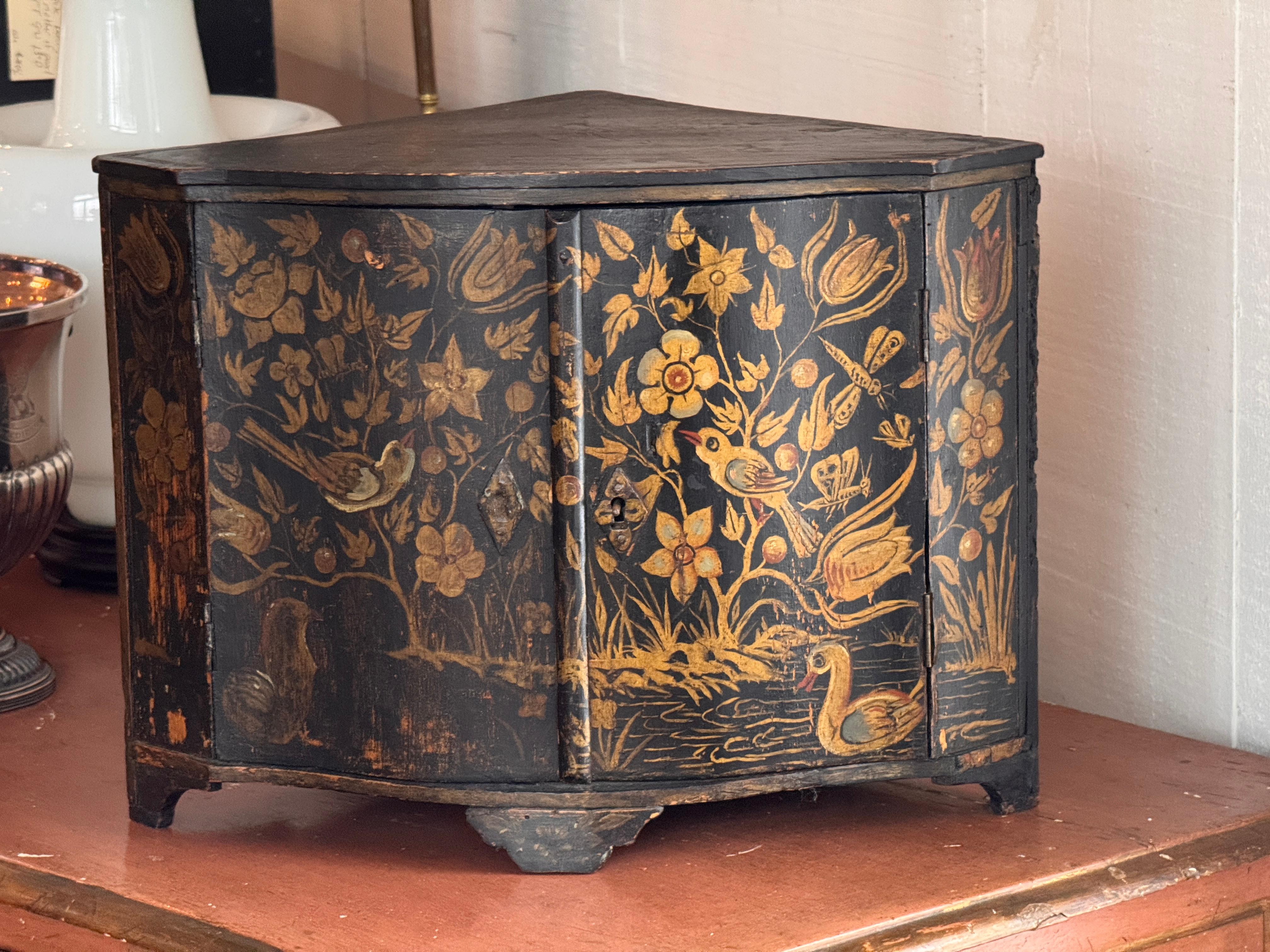 A small painted corner cabinet with a hinged door and floral-and-bird decoration across the front panels. The shaped front sits on short bracket feet, and the interior is simply finished in black. Wear, losses, and age-related surface marks are