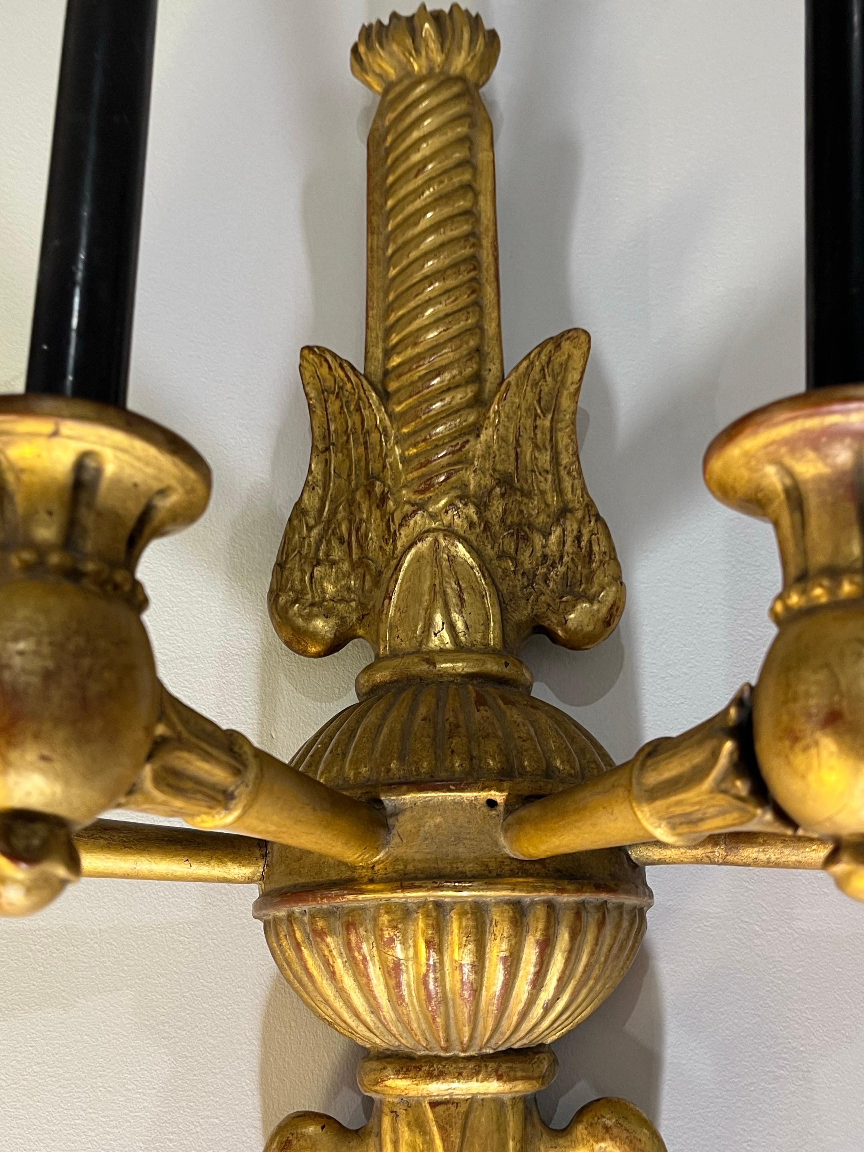 Une belle paire d'appliques murales pour bougies, en bois sculpté et doré, avec des motifs décoratifs typiques de la période Empire, tels que des flammes, des feuilles d'acanthe et des ailes.
Elle comporte au total quatre bras de bougie.

La partie