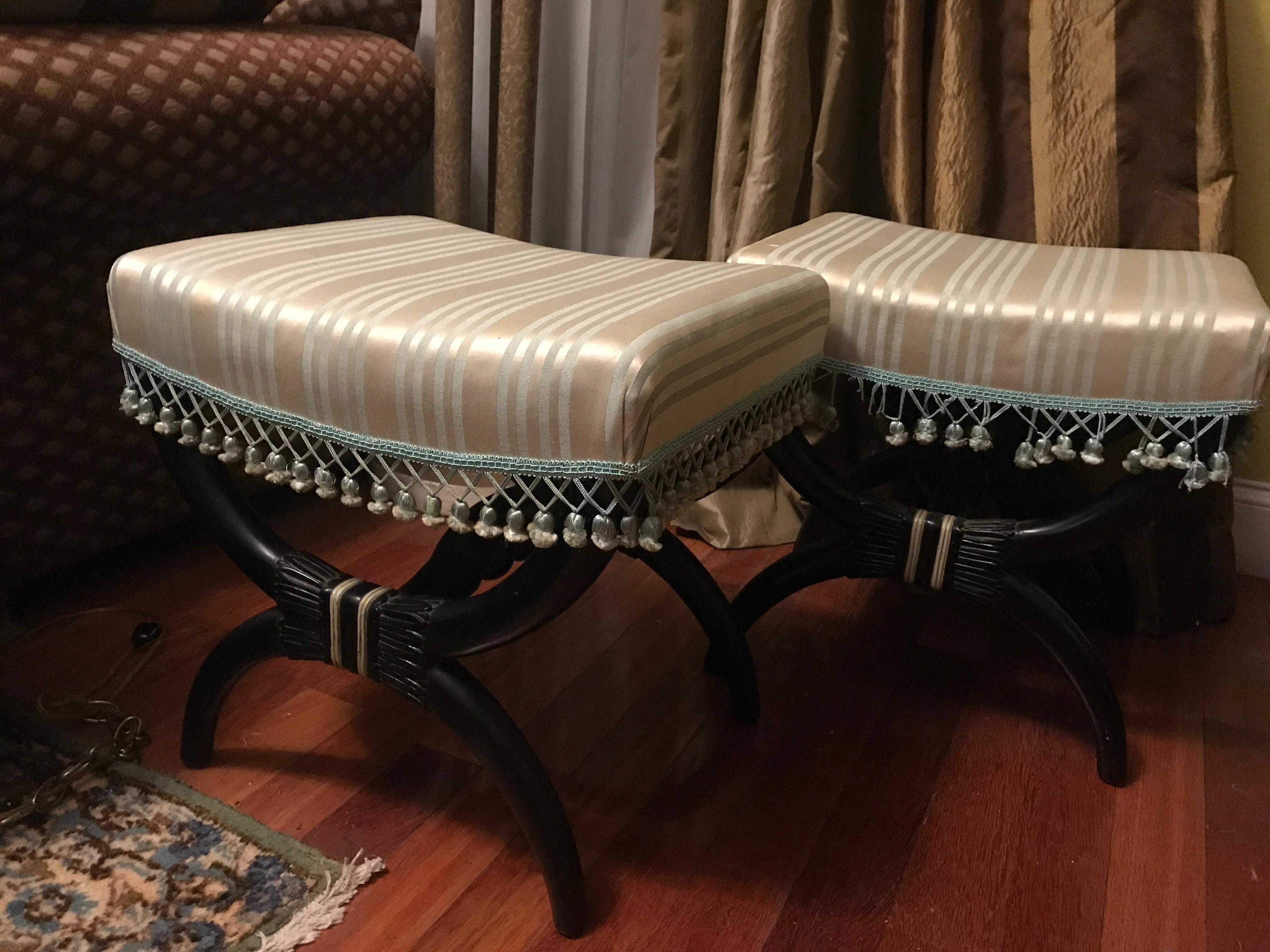 Silk fabric in cerulean blue and egg-shell combines with whimsical trim that accents the deep tone wood of two curule benches in black, a pair in the neoclassical style, ebonized and upholstered in silk. The pair is American or European, late 19th/
