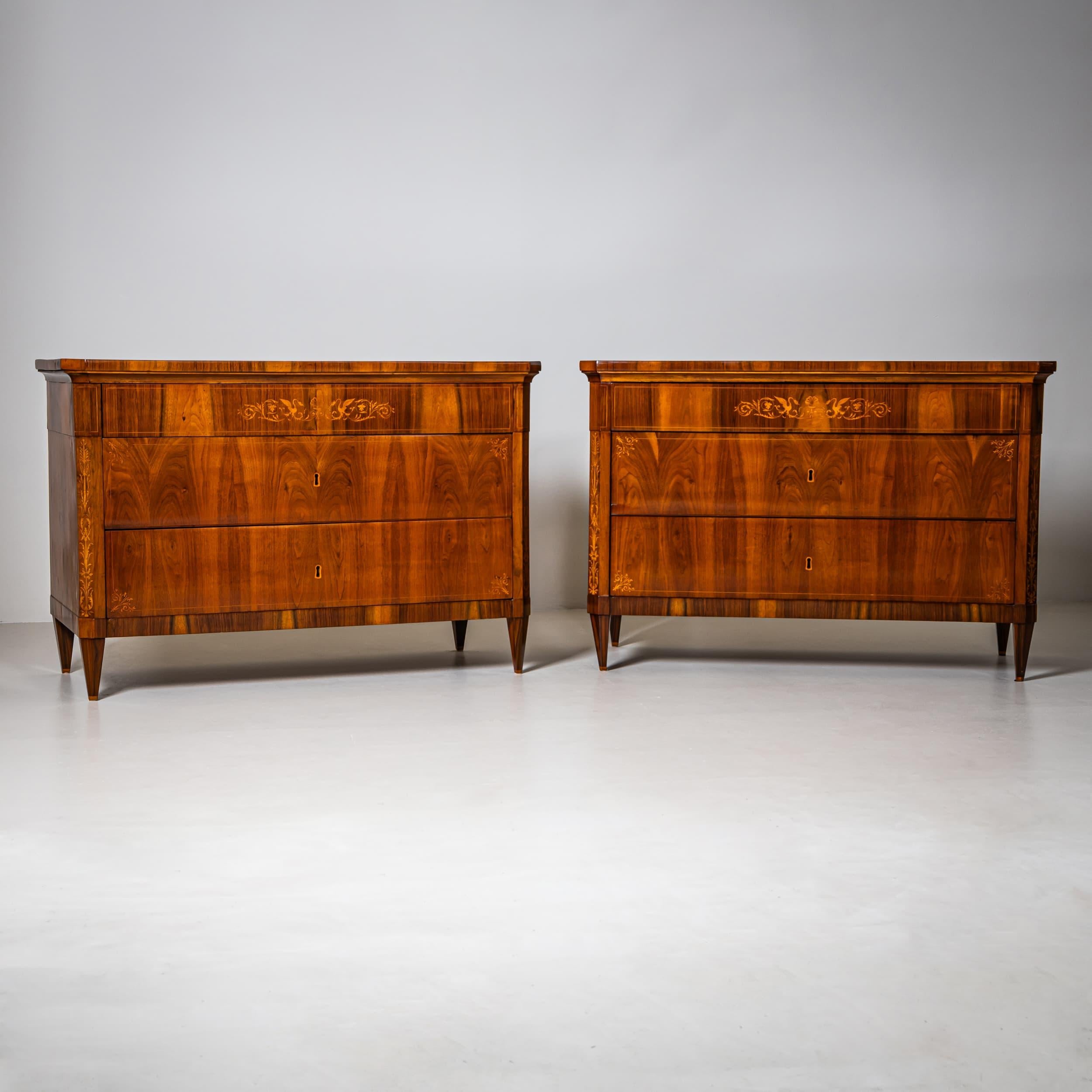 Pair of large Biedermeier chests of drawers with three drawers and chamfered corners. The chests rest on tapered square feet, and the profiled top slightly overhangs. The top drawer is narrow and features decorative inlays of swans and scrolling