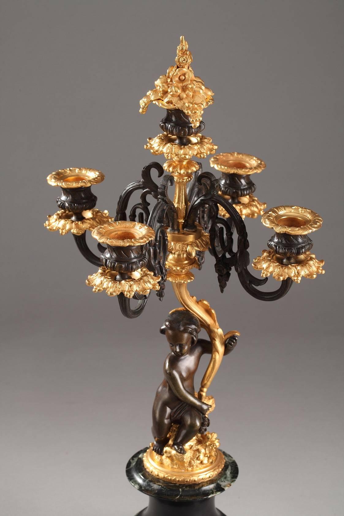 Pair of gilt and patinated bronze candelabras. Each Rocaille-inspired candelabra is ornamented with a putto seated on a rock and holds the stem of the candelabra in his raised arm. The gilded, curved stem raises to support five branches that are