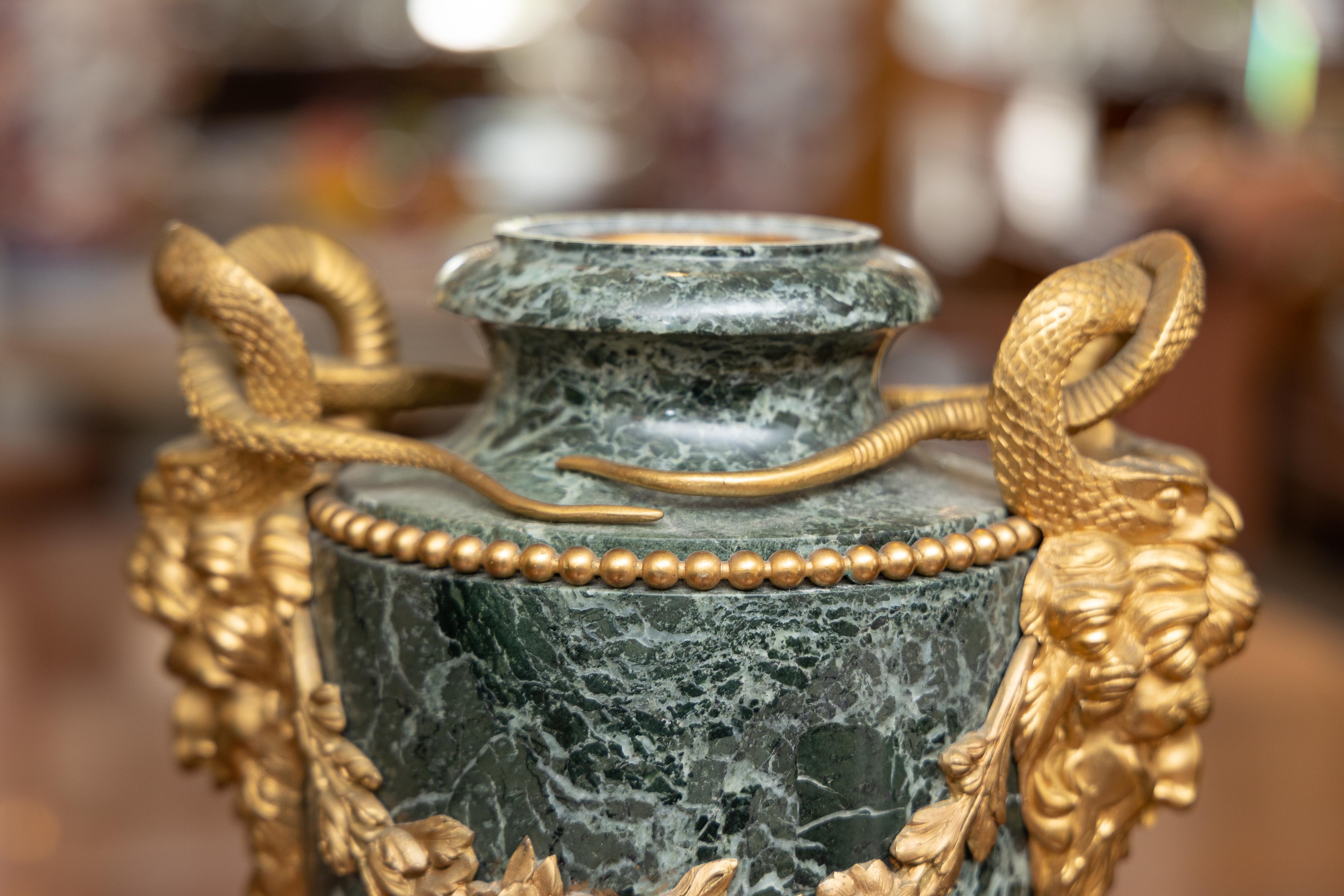 This is a stately pair of French verdi green marble urns. The urns are adorned by gilt bronze reptiles and a pair of male masks joined by a decorative swag, situated on a marble base, 19th century.