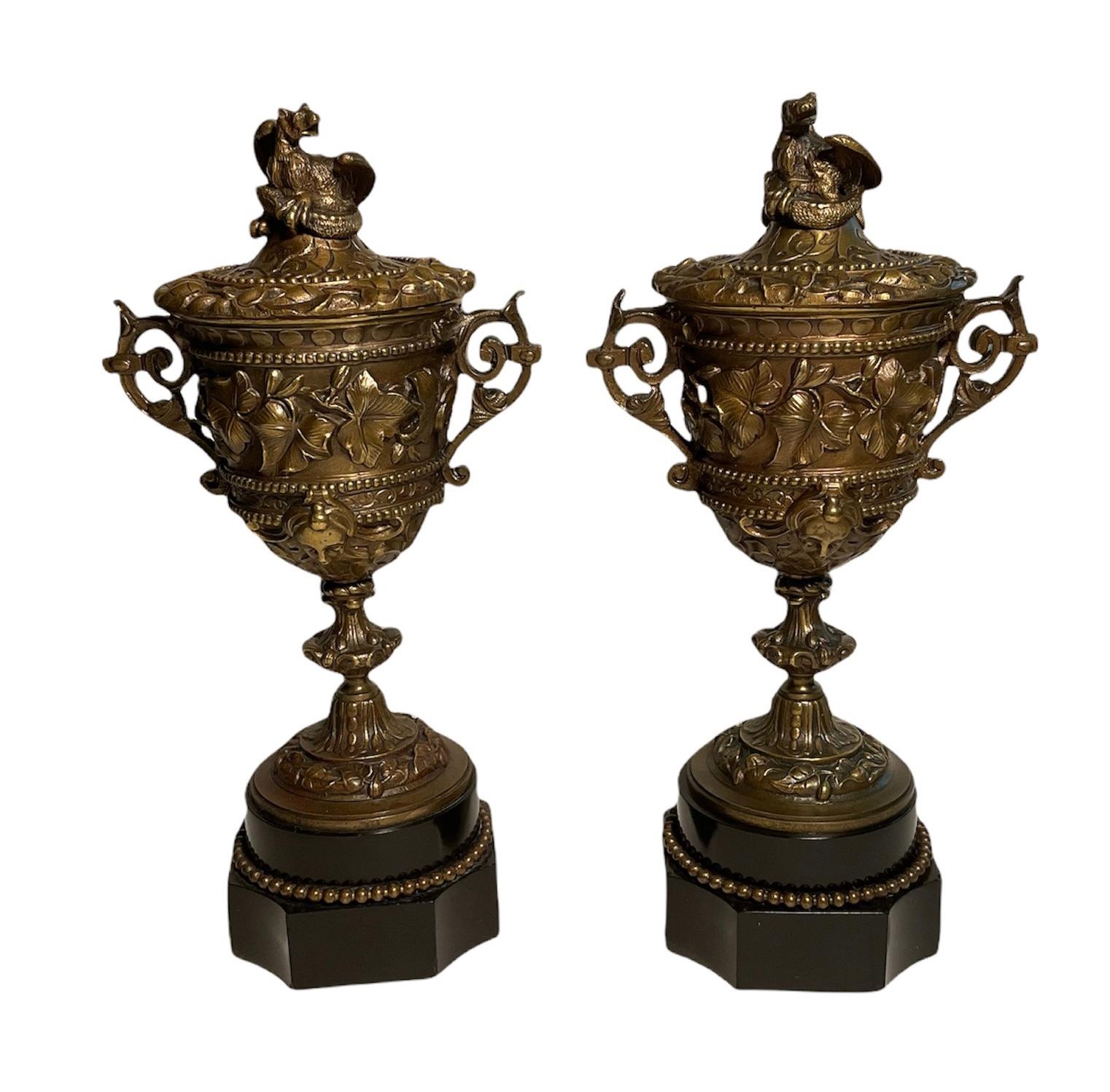 This is a pair of patinated bronze cups with covers. A repousse of ivy leaves garland decorates the center of the cups’ body. Above and below it, there are a chain of beads adorning it. Below them, there is a repousse of a head-face of a woman in