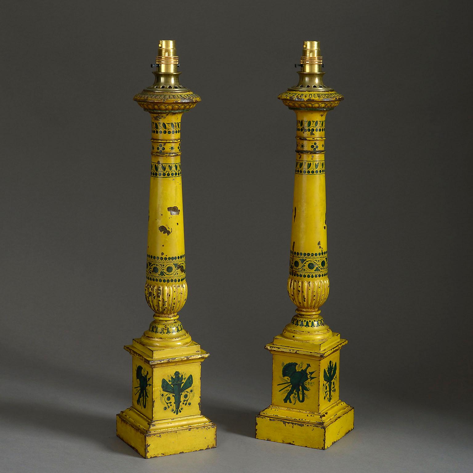 A pair of 19th century tole lamps in the form of columns decorated with bands of stylized flowers and standing on moulded plinth bases decorated with panels of military trophies in shades of petrol blue on a butter yellow ground, wired with silk