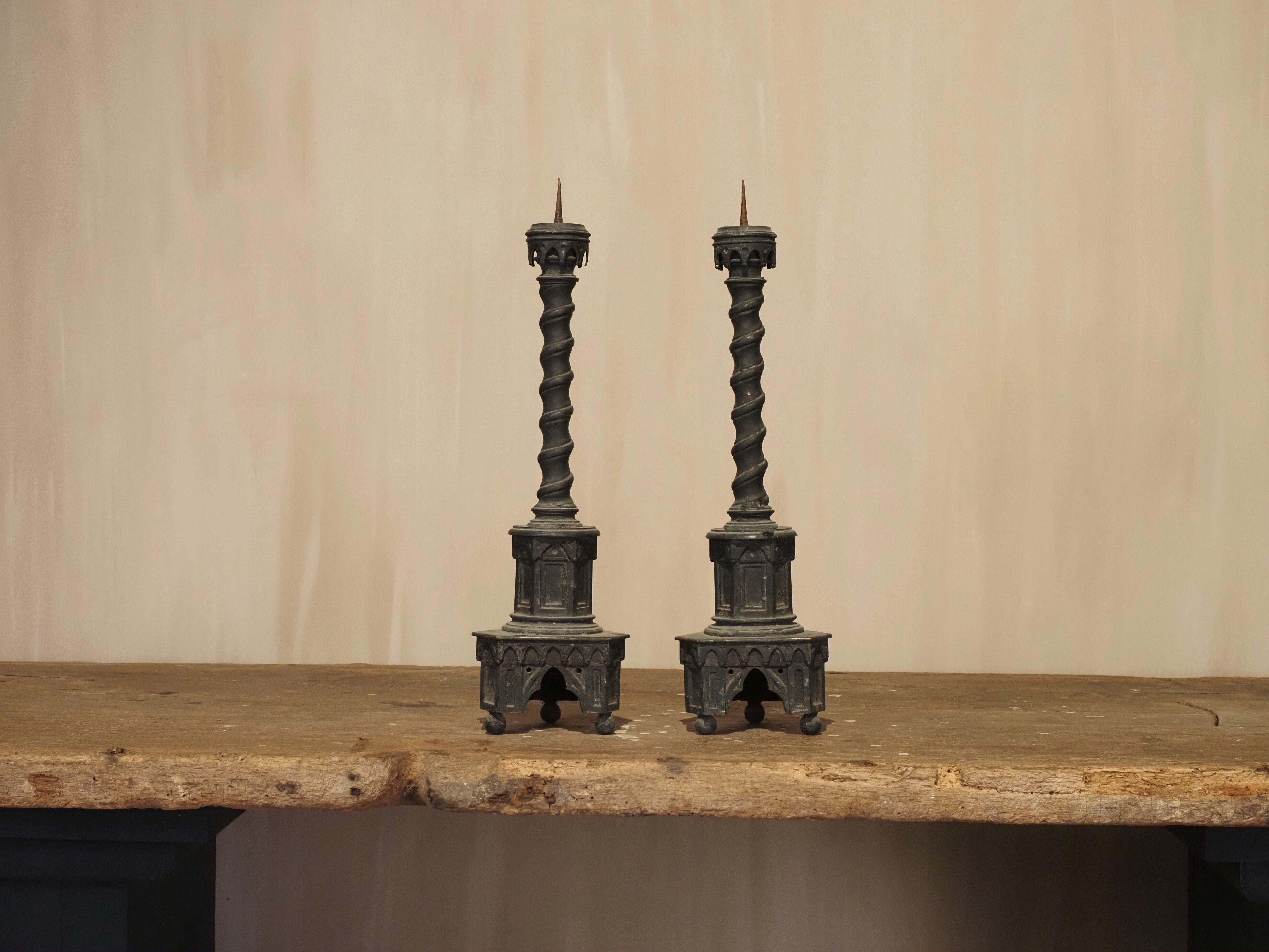 A finely crafted pair of 19th-century neo-Gothic altar candlesticks, cast with a rich dark patina. Each candlestick stands on a square base with pointed arch decoration, raised on three small feet, and features an openwork niche on each side. The