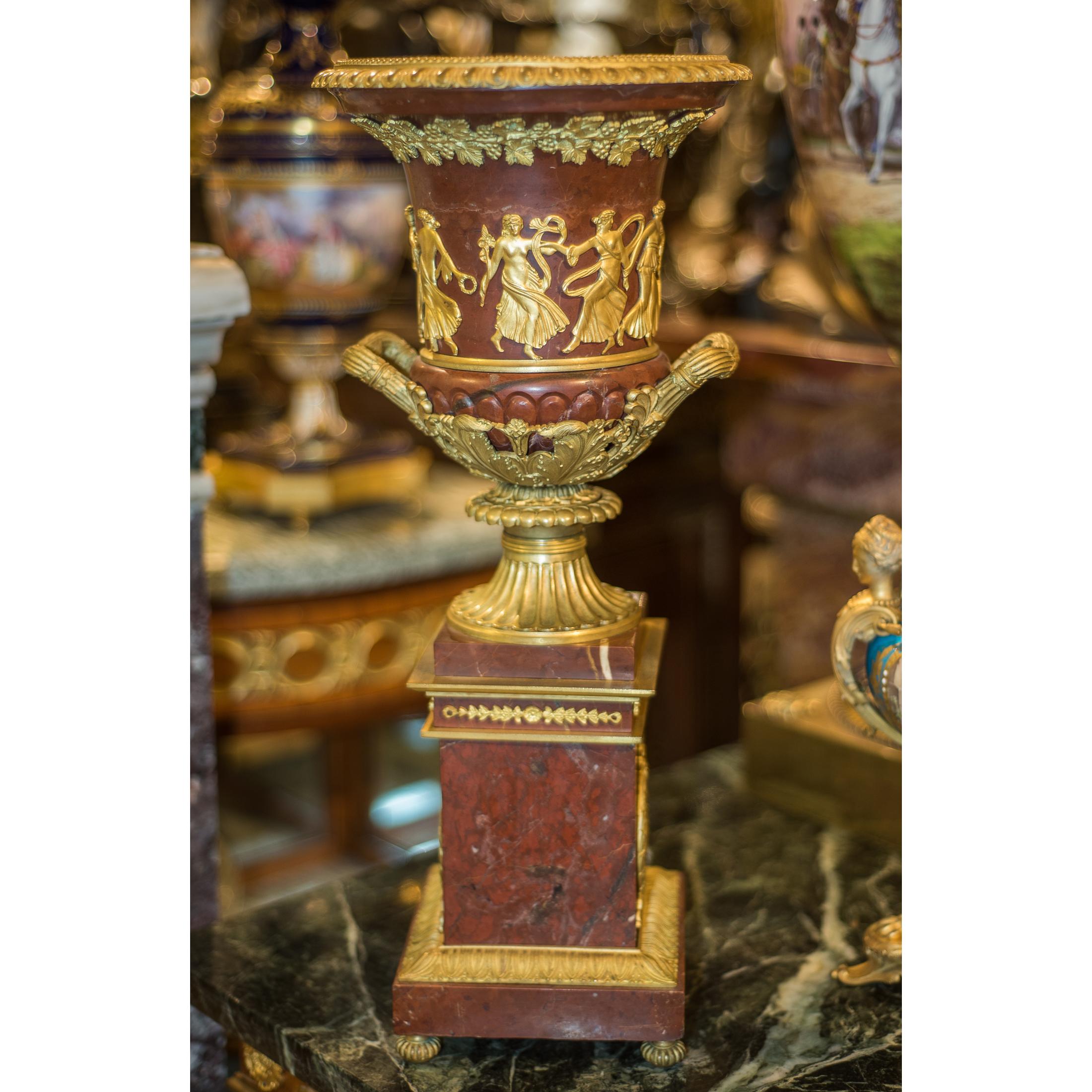 A fine quality pair of ormolu-mounted rouge marble urns featuring neoclassical dancers. A fine pair of French Regency gilt bronze mounted rouge marble urns featuring a neoclassical frieze of dancers on the surface with a column base bodice adorned