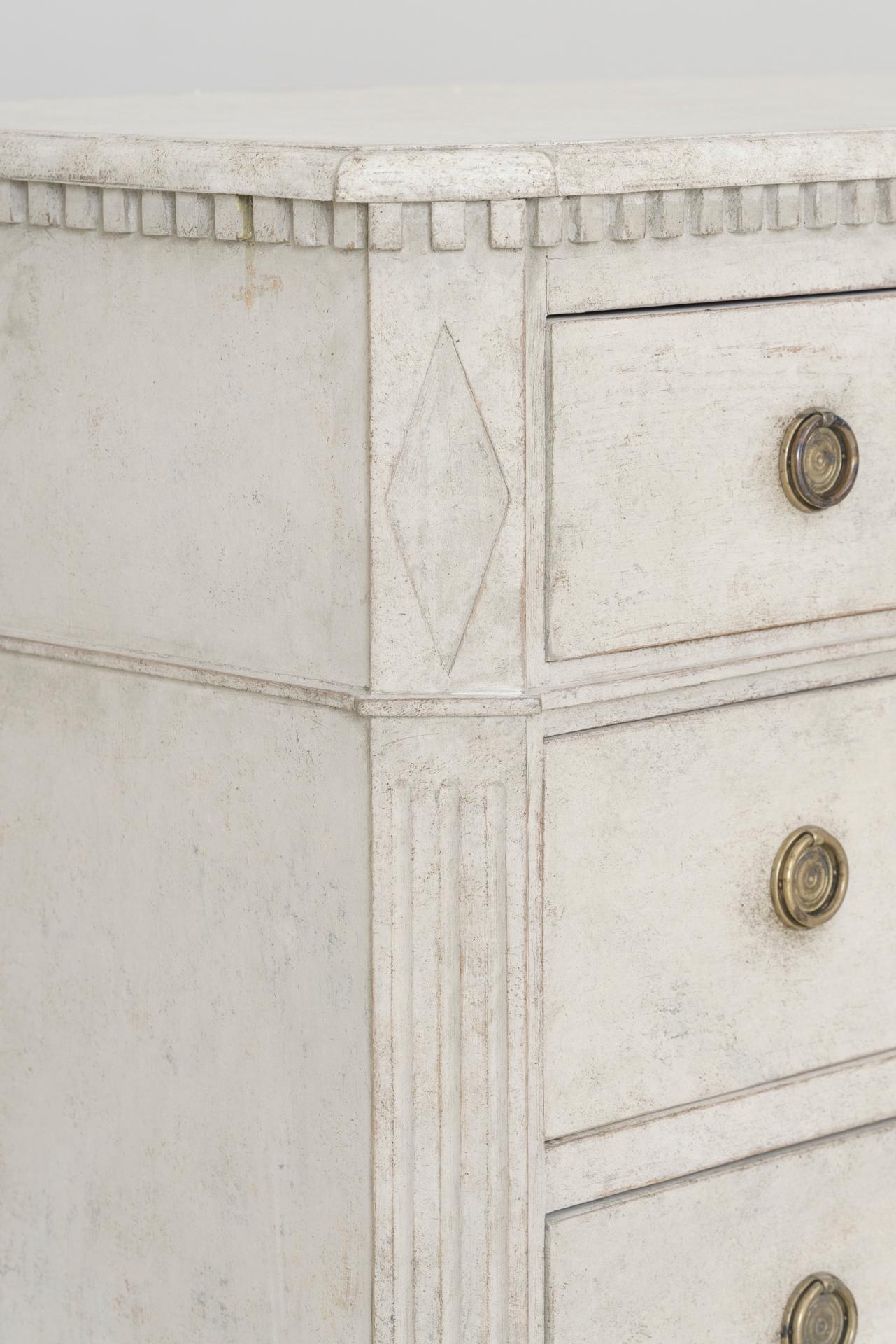 A pair of Swedish commodes in the Gustavian style with dentil molding around the top and lozenge detail on the corners. Chalky ivory paint with a gray hue. Three large drawers, brass hardware, canted and fluted corner posts, and tapered and fluted