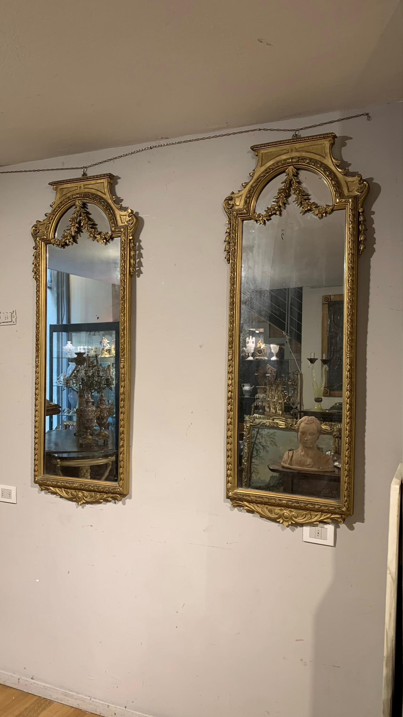 Splendid pair of mirrors in neoclassical style in carved and gilded wood with pure gold leaf. The mirrors are decorated with delicate vegetal racemes that extend up to the upper part, where they join in a decorative garland that also passes above